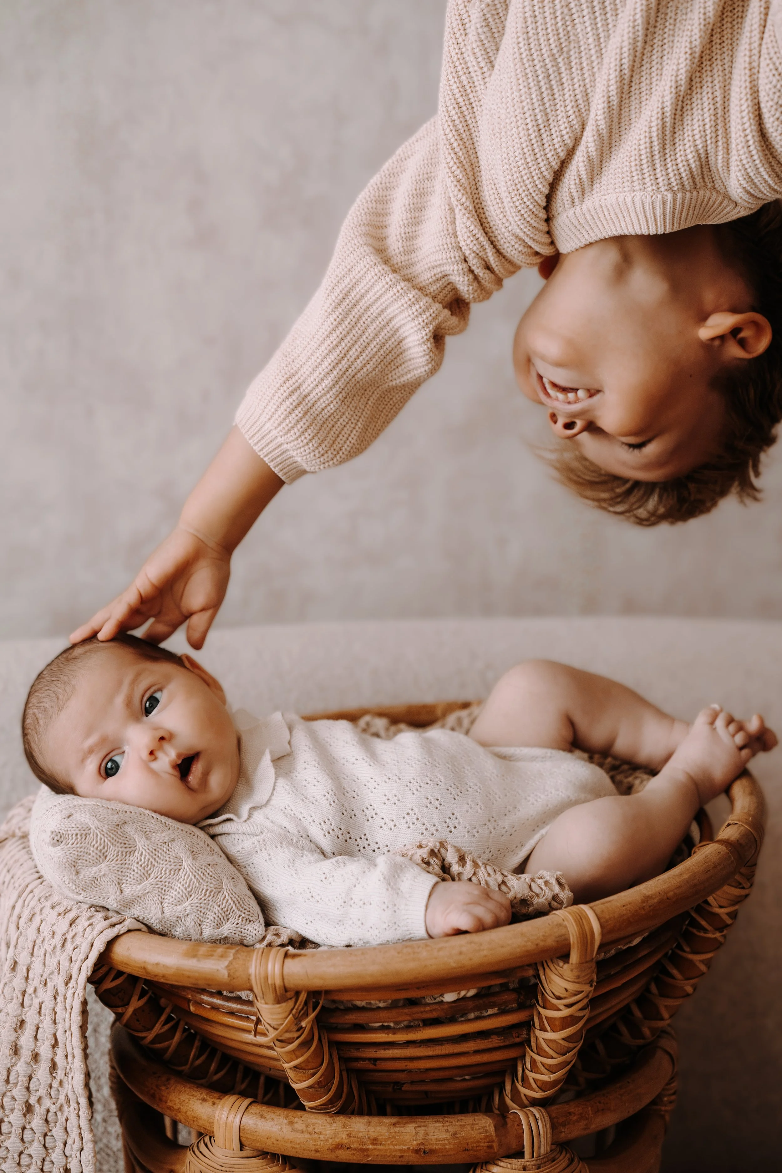 Zwei Babys in einem Korb, eines liegt und schaut in die Kamera, das andere sitzt und lacht, während eine erwachsene Person sie berührt.