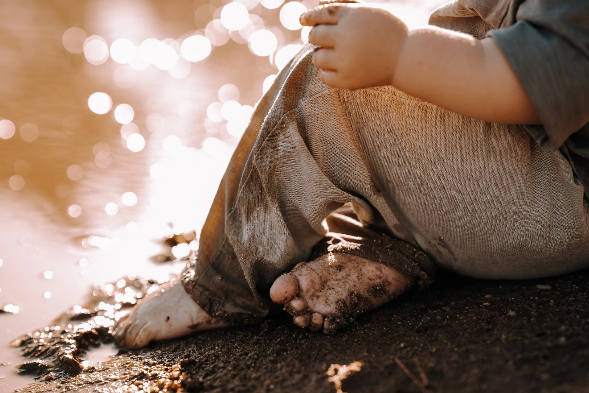Nahaufnahme eines Menschen, der mit bloßen Händen und einem Werkzeug Sand am Wasserufer bewegt. Das Bild ist warm beleuchtet mit Bokeh-Lichteffekten im Hintergrund.