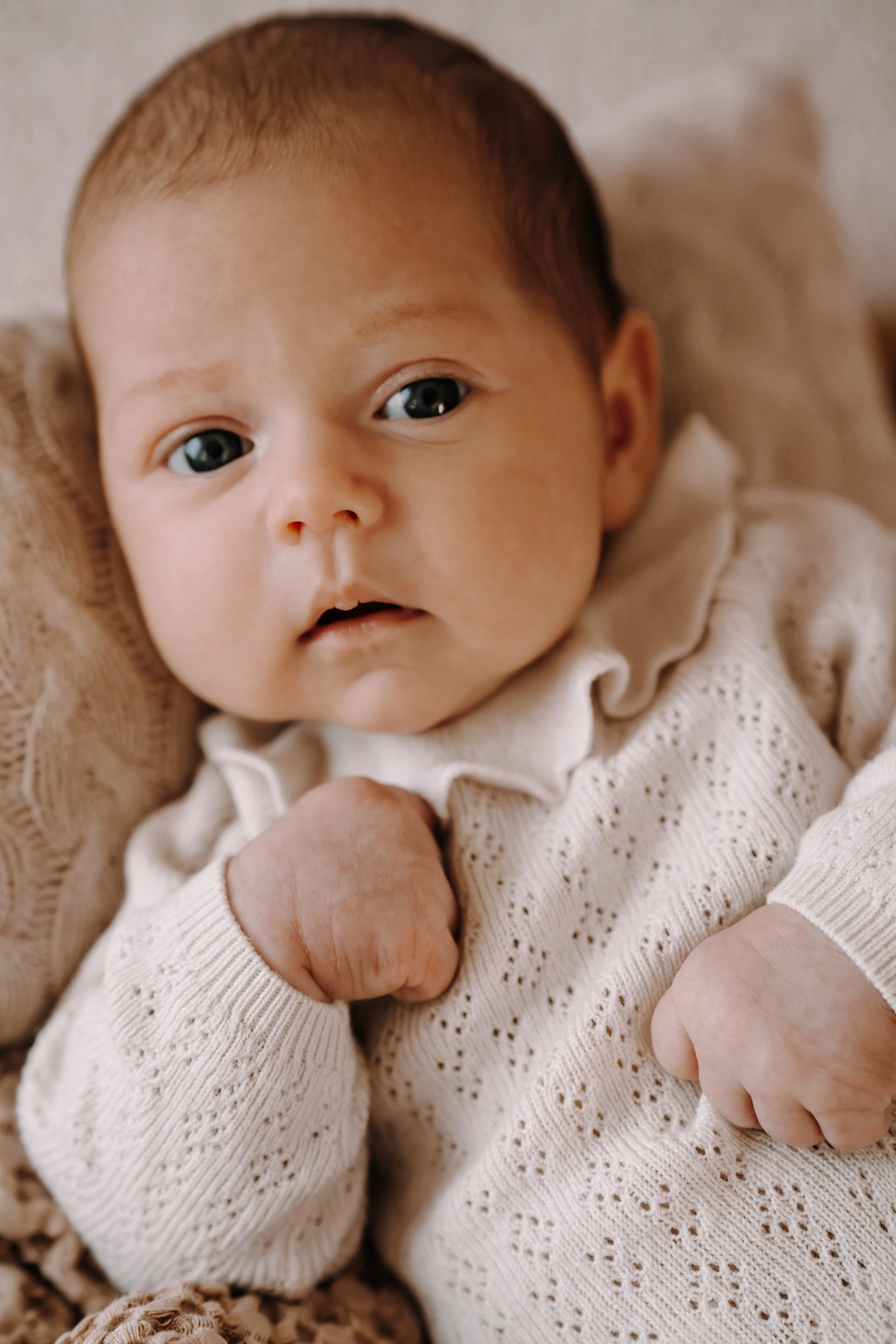 Nahaufnahme eines neugeborenen Babys in einem weißen Strickpullover, das auf einem weichen, beige-braunen Untergrund liegt. Das Baby schaut direkt in die Kamera, die rechte Hand ist zu einer Faust geballt, die linke Hand ist nur halb sichtbar.