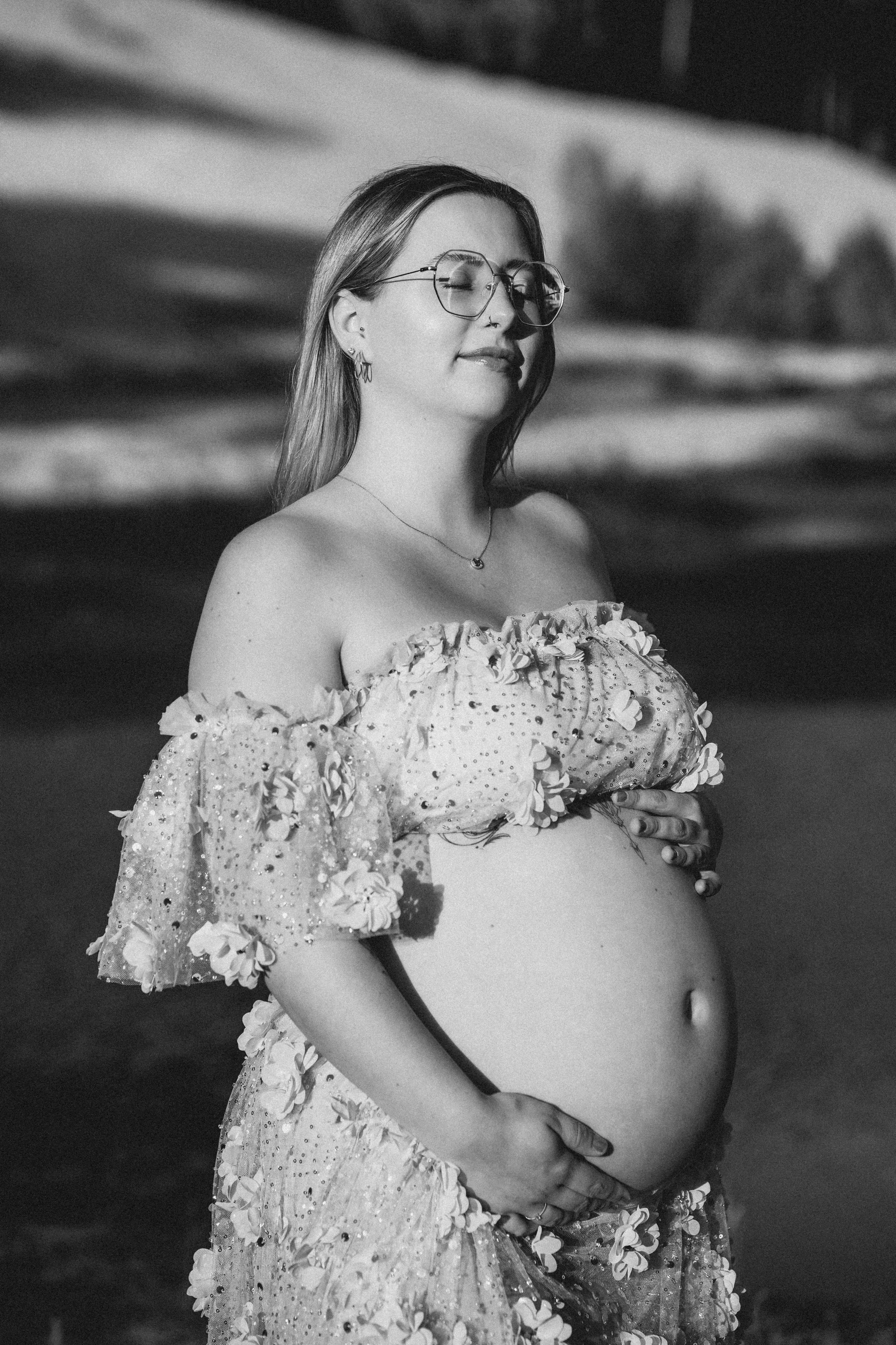 Schwangere Frau in einem floral verzierten Kleid, die auf einer Wiese steht und die Sonne im Gesicht spürt, in Schwarz-Weiß.