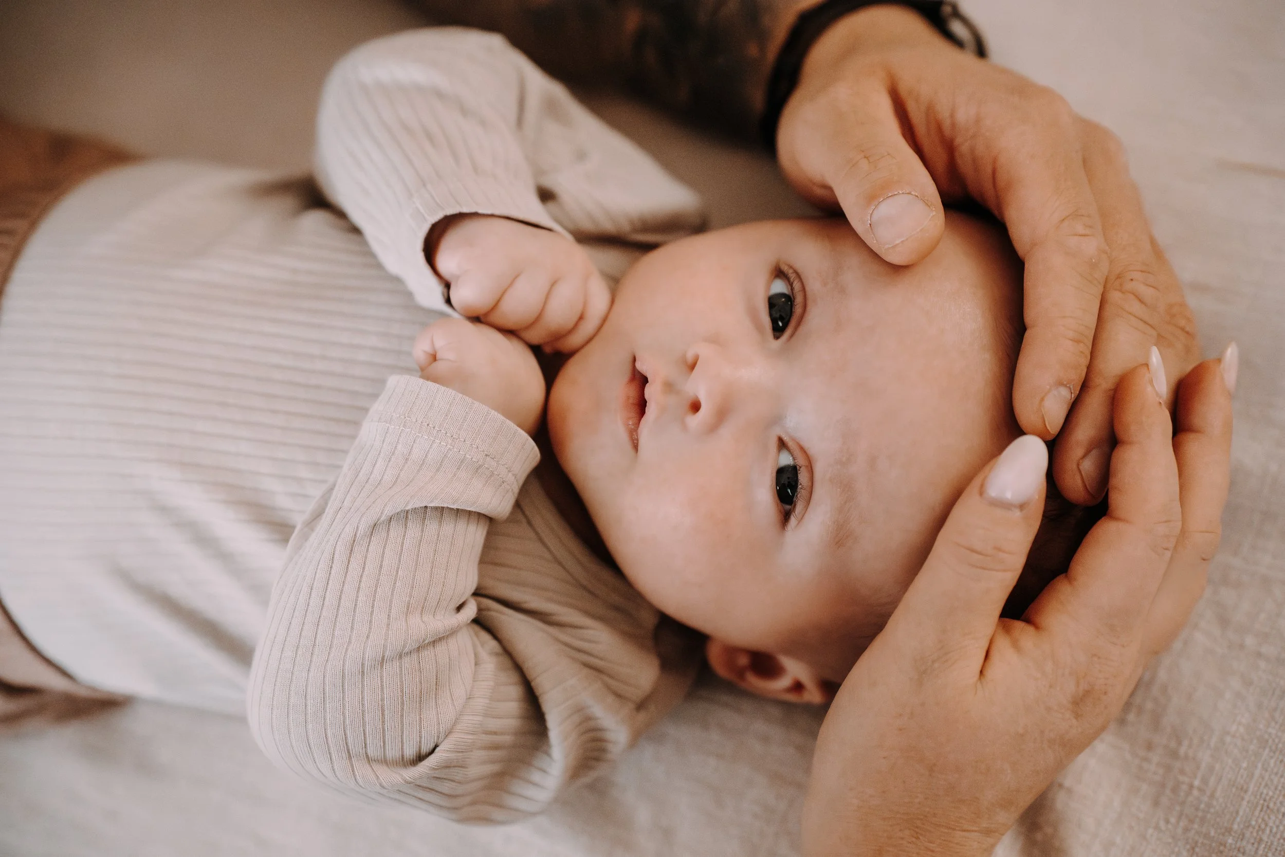 Ein Baby liegt auf einem weichen, hellen Tuch und wird von einer erwachsenen Person liebevoll gehalten. Die Person streichelt vorsichtig den Kopf des Babys, das in einem hellen Kleid mit langen Ärmeln gekleidet ist.