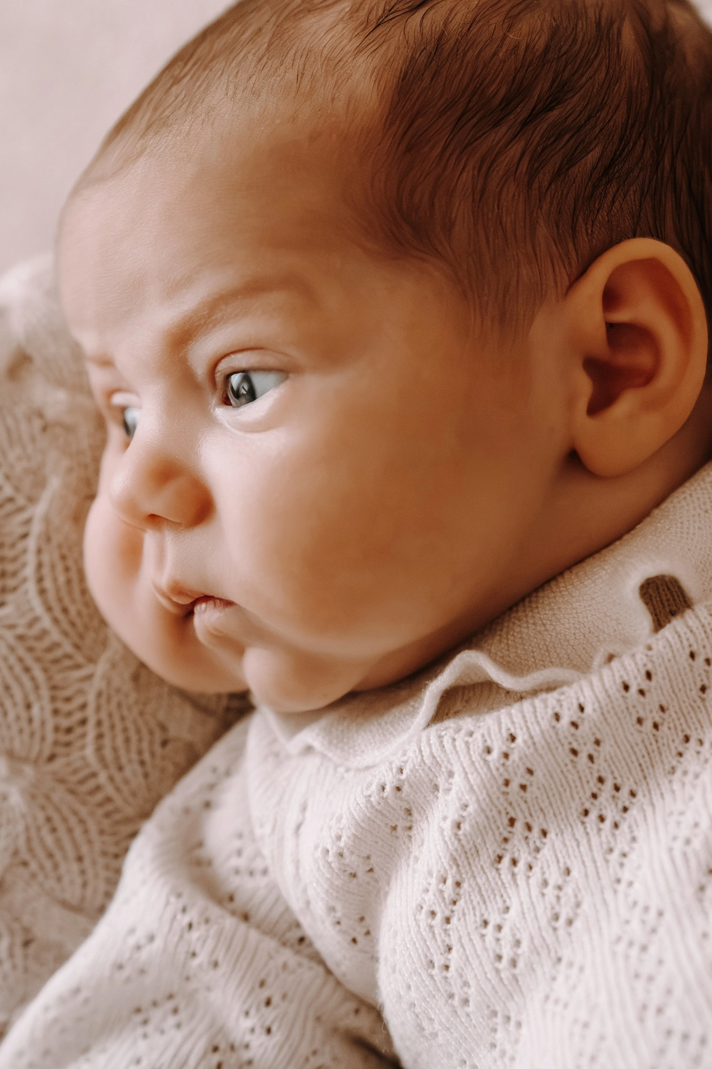 Nahaufnahme eines Babys mit hellbraunen Haaren, das in einem weißen, gestrickten Kleid liegt.