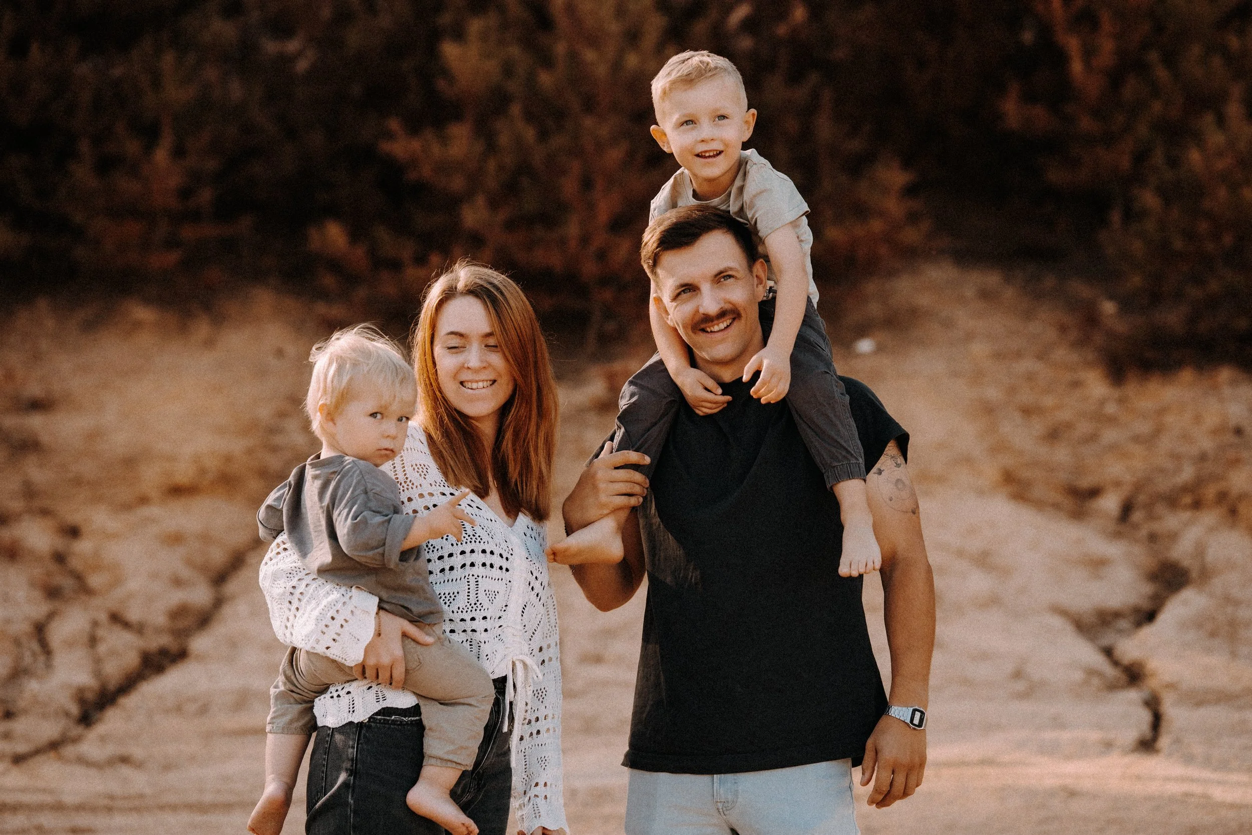 Familie mit vier Mitgliedern beim Spaziergang im Sonnenschein, der Vater trägt ein Kind auf dem Rücken, die Mutter hält ein weiteres Kind, alle lachen und genießen die Natur im Freien.