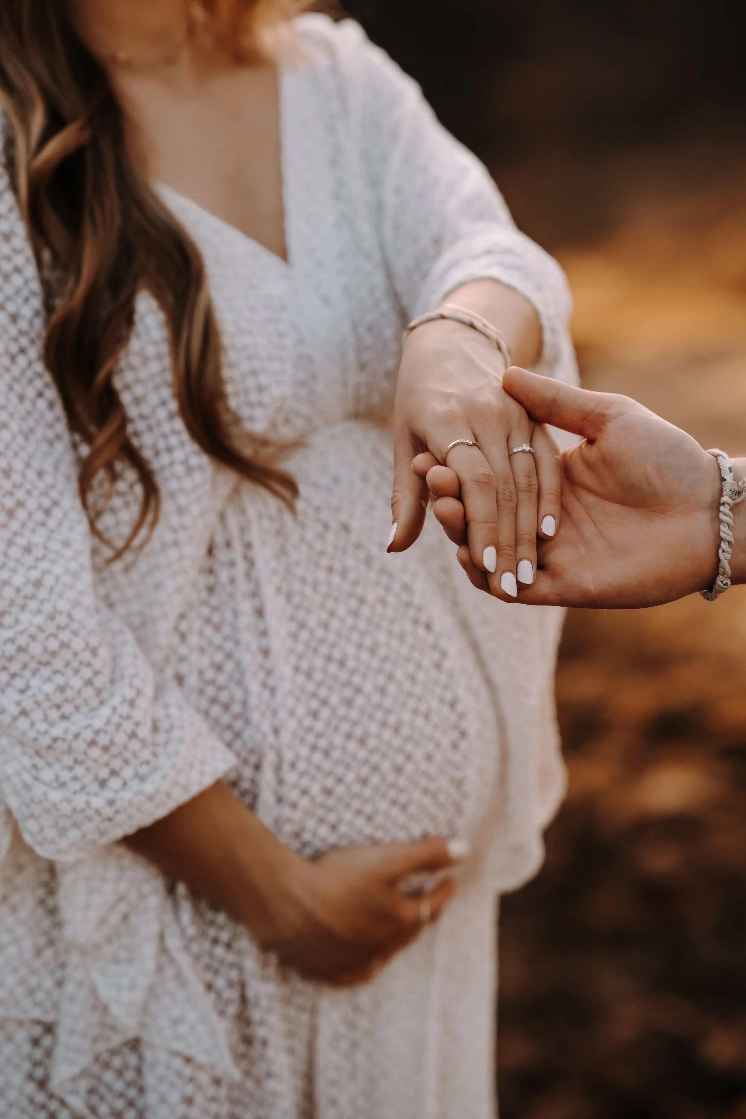 Ein schwangere Frau in einem weißen Kleid hält ihre Hände mit einem anderen Paar in der Nähe. Die Hände tragen Ringe und Armbänder, im Hintergrund ist eine natürliche, warme Beleuchtung.