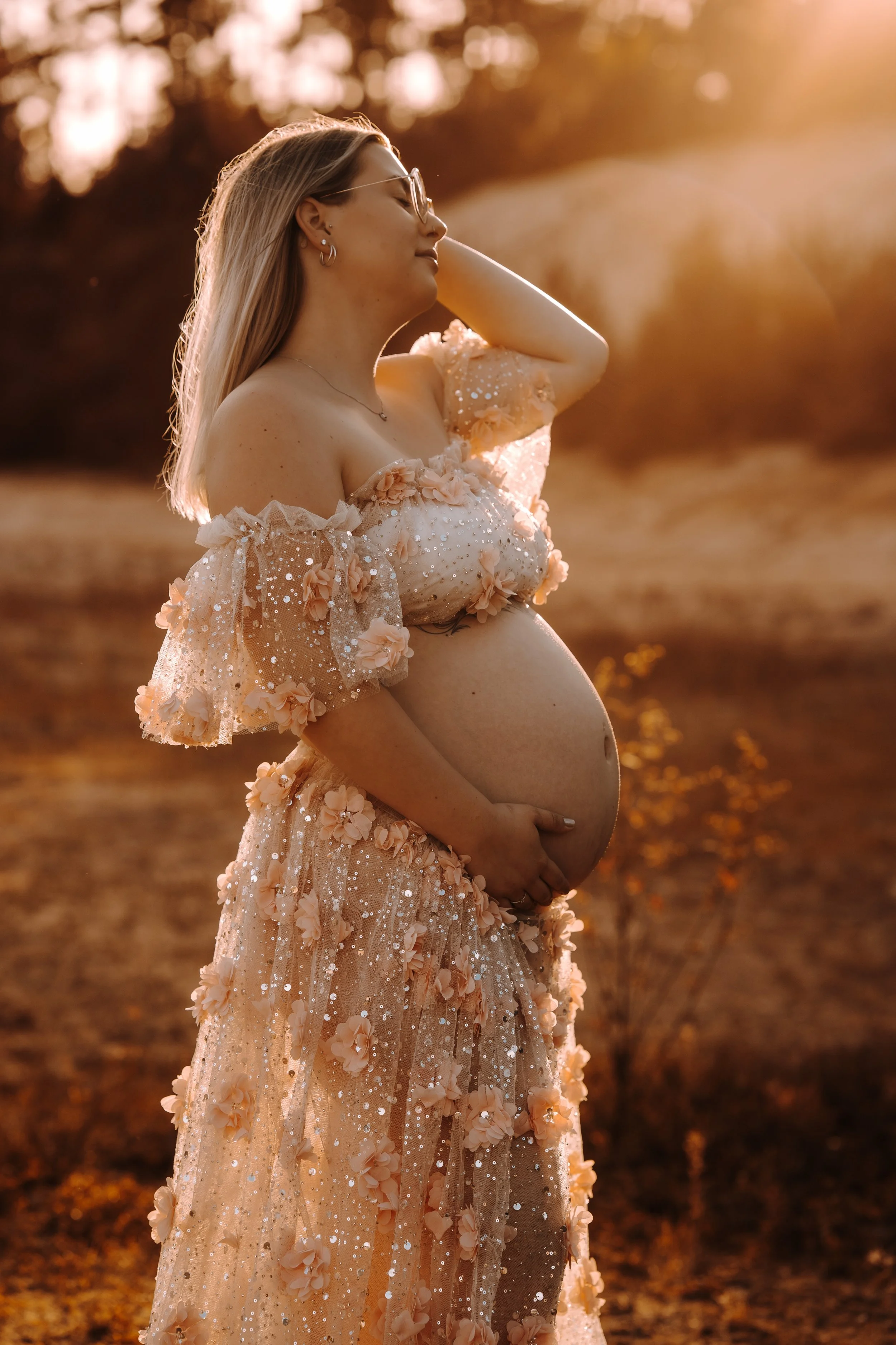 Eine schwangere Frau in einem cremefarbenen, blomsterbestickten Kleid im Sonnenuntergang. Sie steht mit geschlossenen Augen und legt ihre Hand auf den Bauch, im Hintergrund sind Bäume und goldenes Licht.