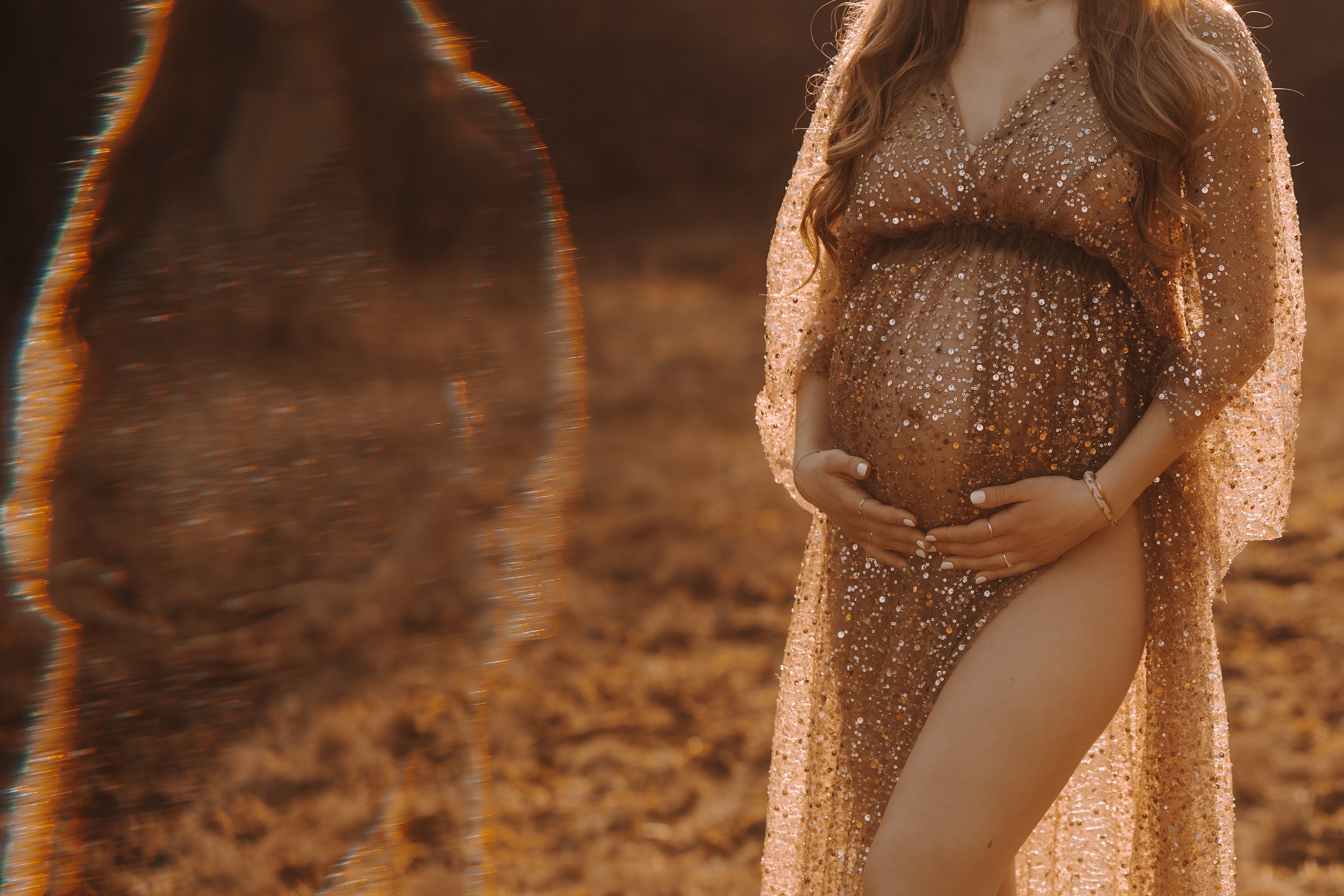 Eine schwangere Frau in einem glitzernden, durchsichtigen Kleid, die ihre Hände auf ihren Bauch legt, im Sonnenuntergang im Freien.