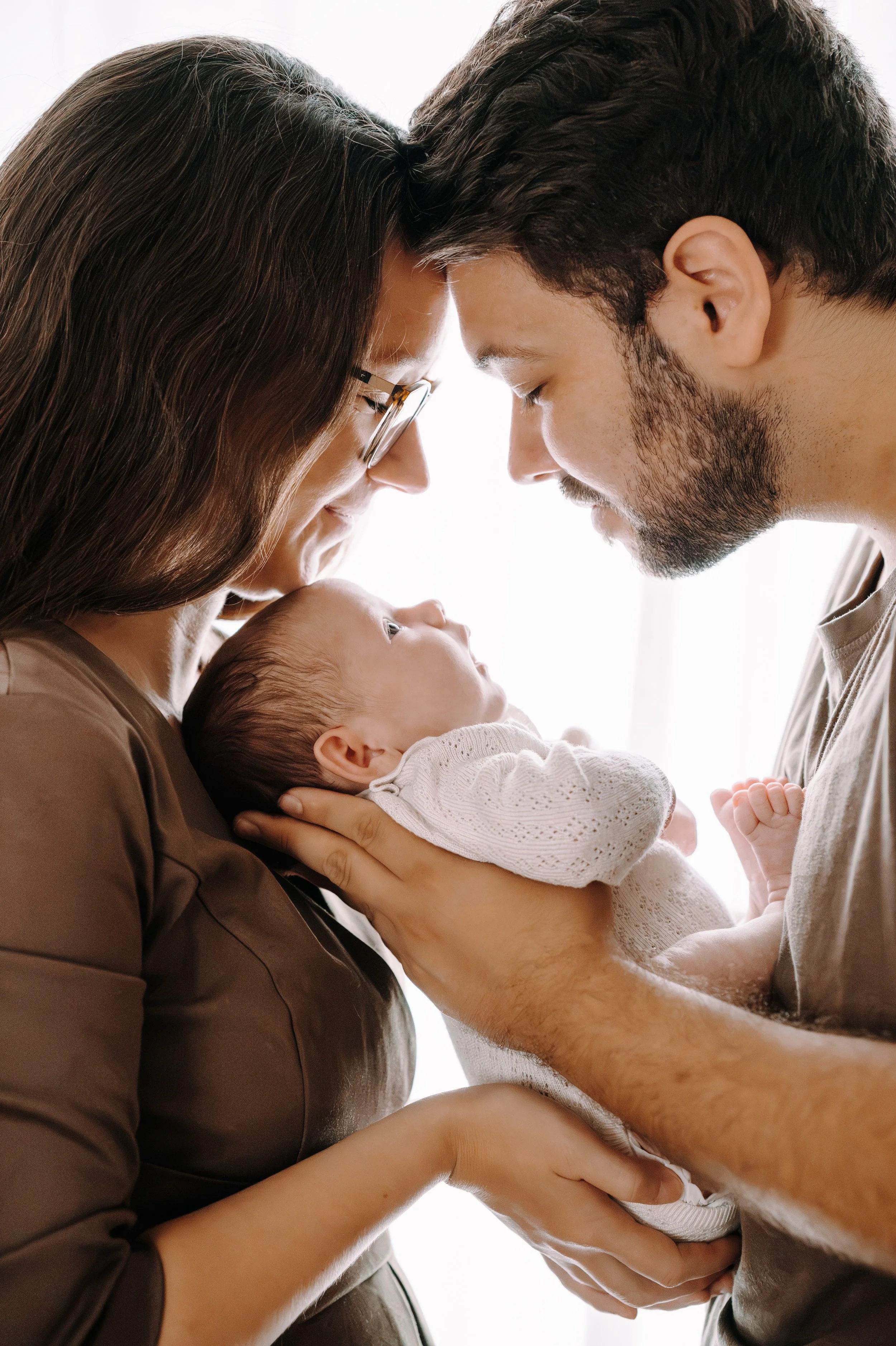 Ein Baby wird von zwei Erwachsenen gehalten und liebevoll berührt, sie berühren die Stirn des Babys mit ihren Stirnen, in einem hell erleuchteten Raum.