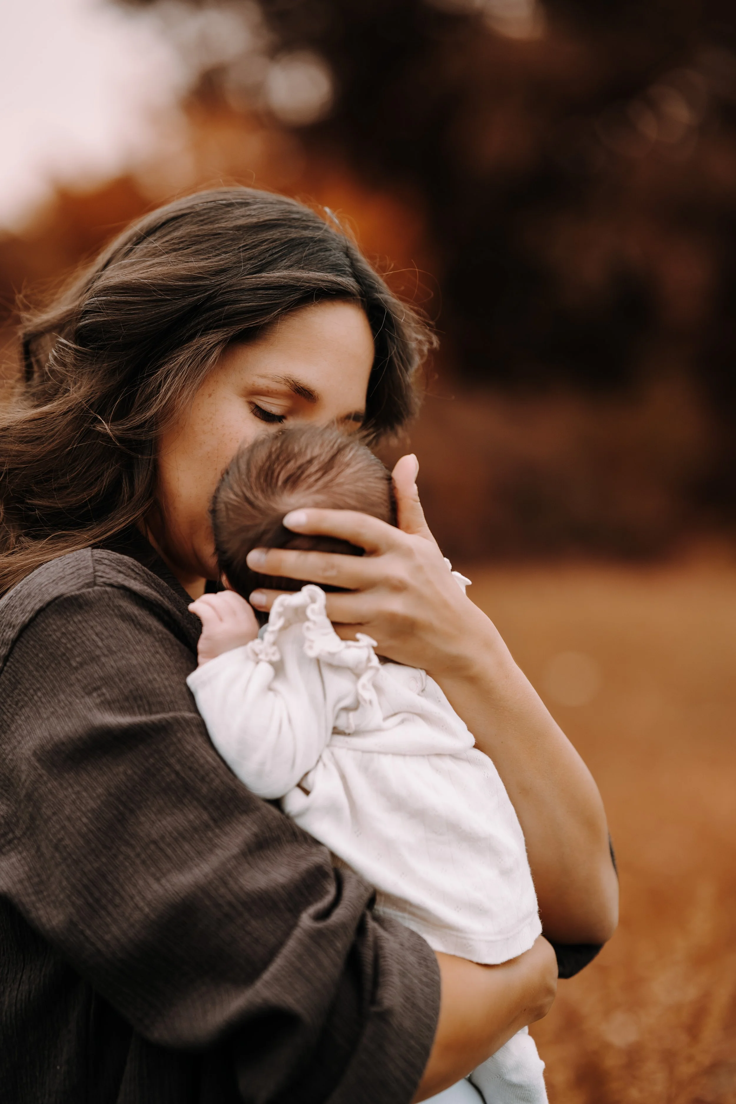 Eine Frau hält ein Baby liebevoll im Arm und küsst es auf die Stirn, im Hintergrund sind herbstliche Bäume.