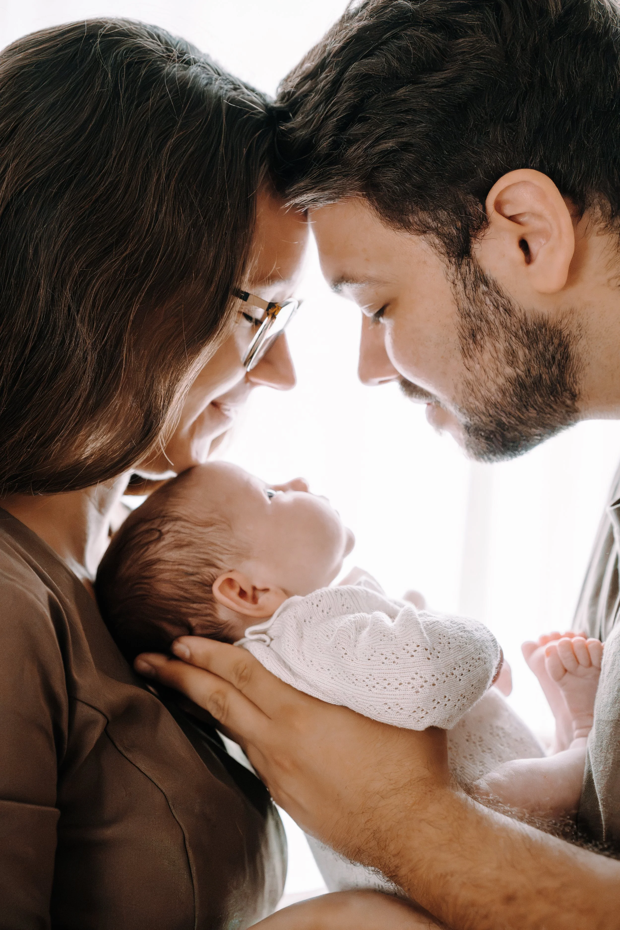 Ein Mann hält ein neugeborenes Baby in den Armen und eine Frau mit Brille berührt mit ihren Stirnen den Mann, während sie liebevoll auf das Baby schauen.