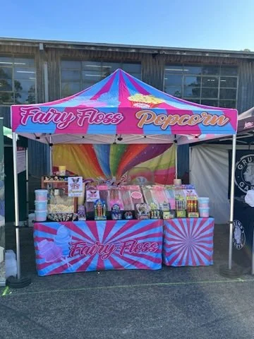 Colorful carnival snack stall selling fairy floss and popcorn under a striped pink, blue, and yellow canopy with rainbow backdrop.