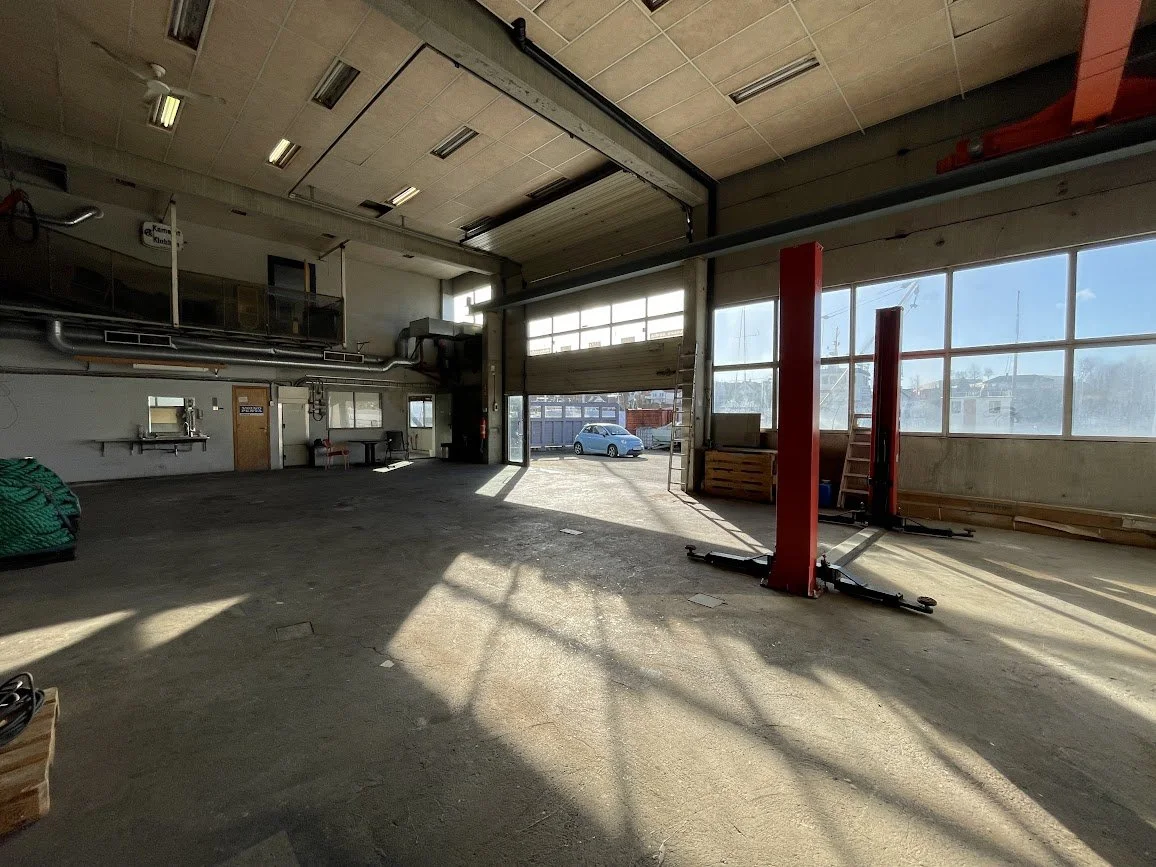 Innendørs bilverksted med en åpen garasjedør, sollys som skinner inn, og en bil parkert utenfor.