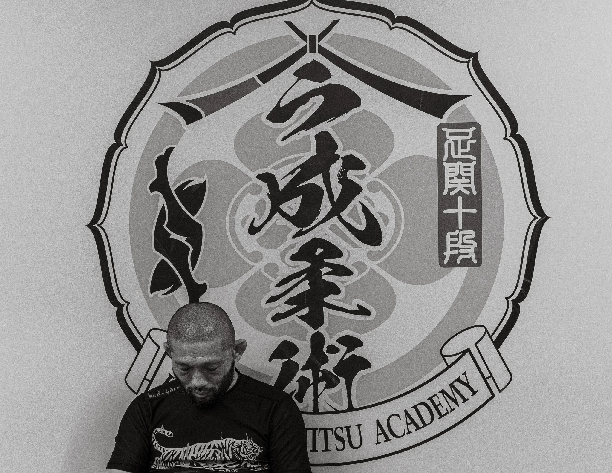 Masakazu Imanari seated beneath the Imanari Jiu-Jitsu Academy emblem inside the Tokyo gym