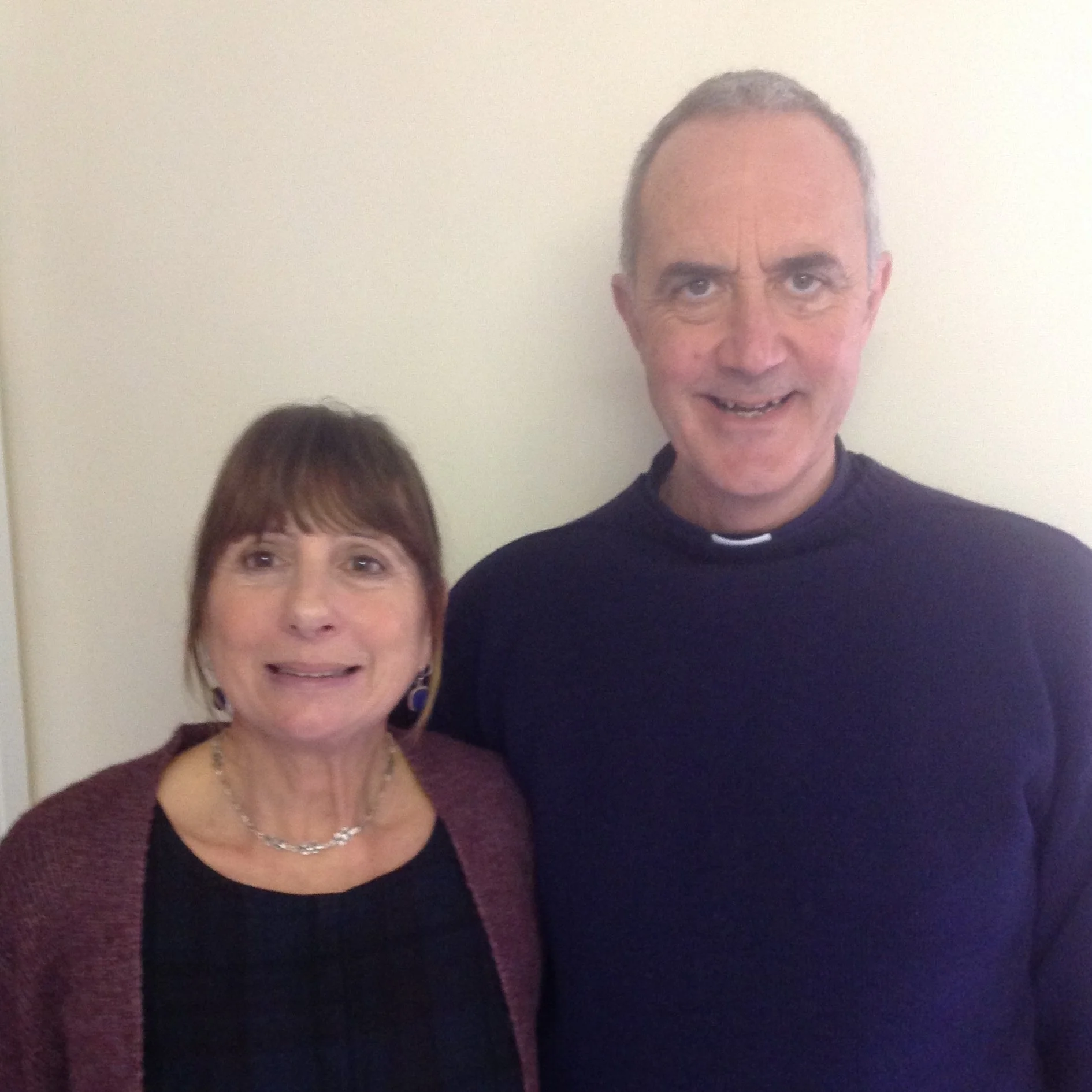 A woman with brown hair and a necklace standing next to a man in clerical attire, both smiling, with a plain white wall in the background.