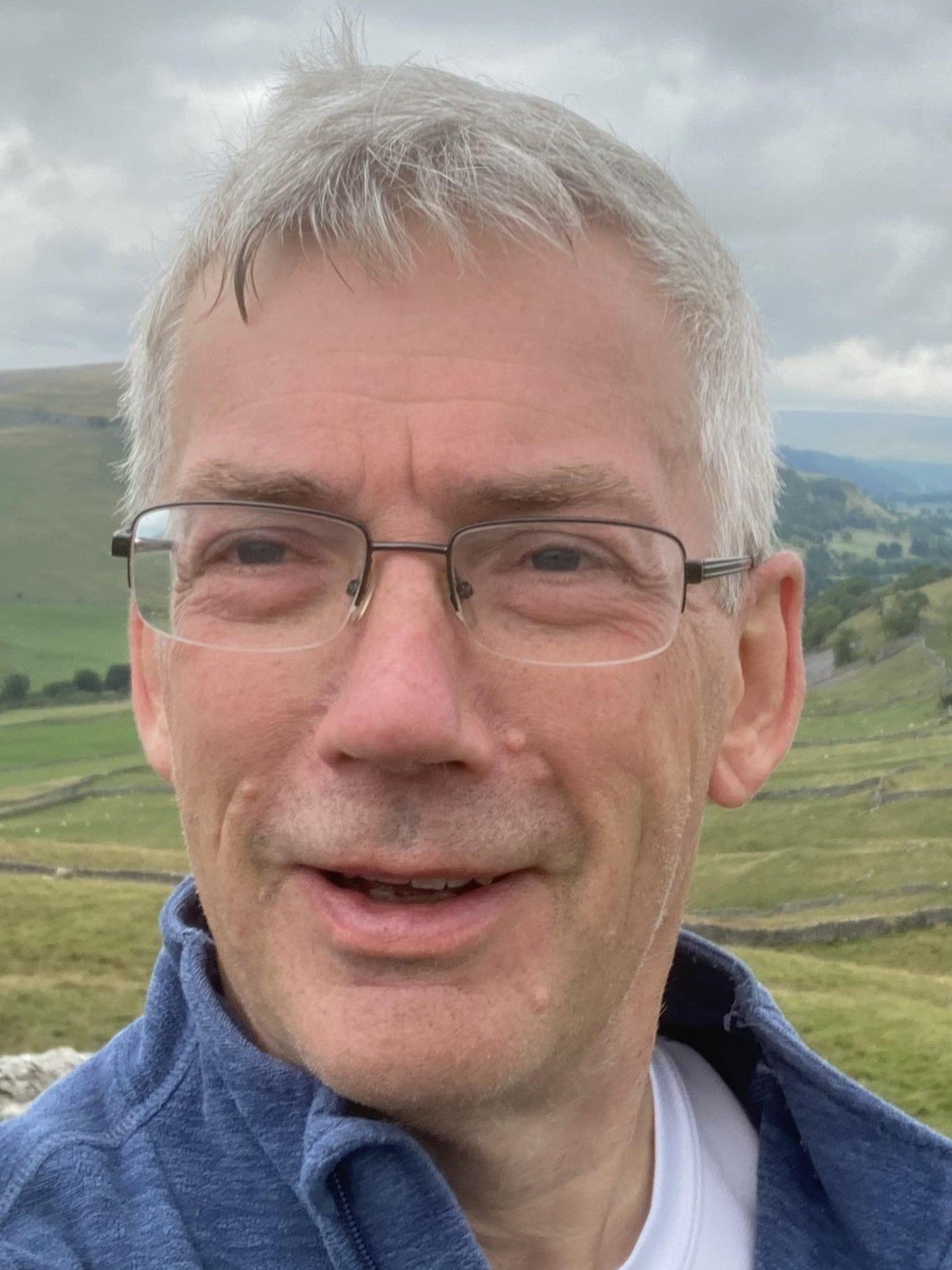 Close-up of a smiling middle-aged man with gray hair, glasses, and a blue shirt, standing outdoors with countryside hills and cloudy sky in the background.