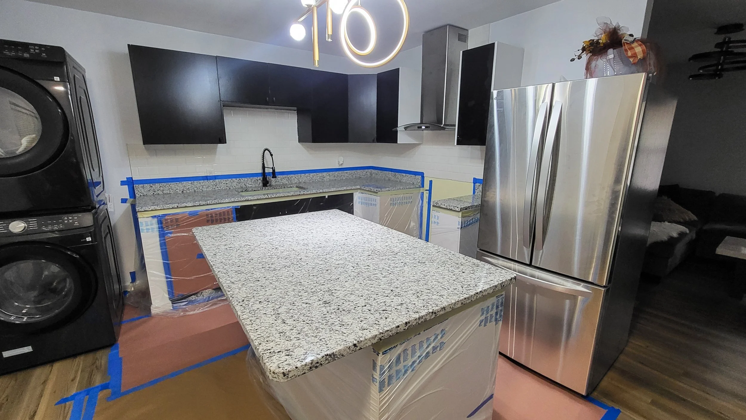 Kitchen with black cabinets, granite countertops, stainless steel refrigerator, black washing machine and dryer, island with granite top, white subway tile backsplash, and a hanging chandelier in the shape of a number 10, decorated with autumn flowers.