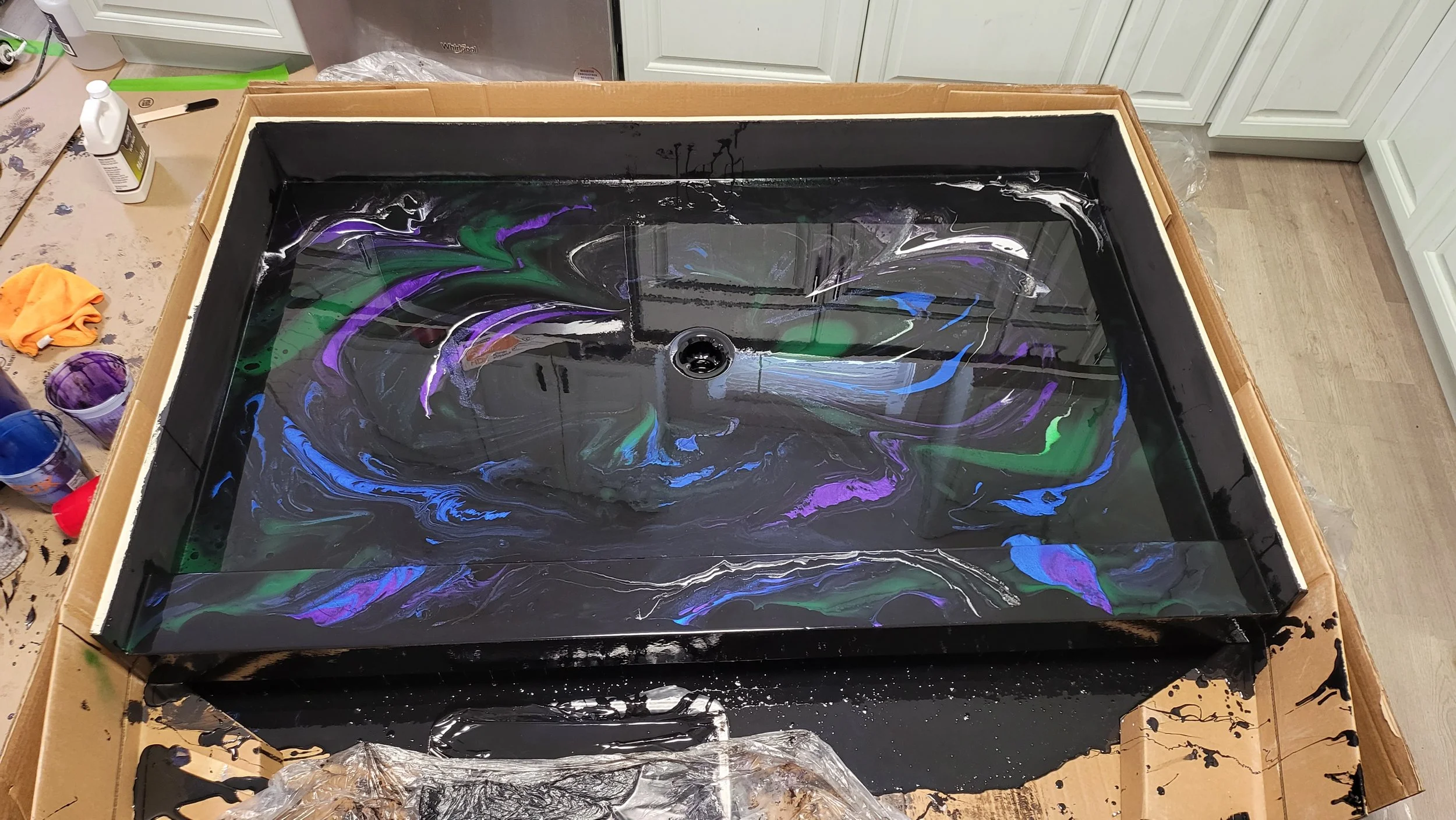 A black epoxy shower pan with swirling patterns of purple, green, blue, and white , placed on a cardboard-covered surface in a kitchen or workshop setting.
