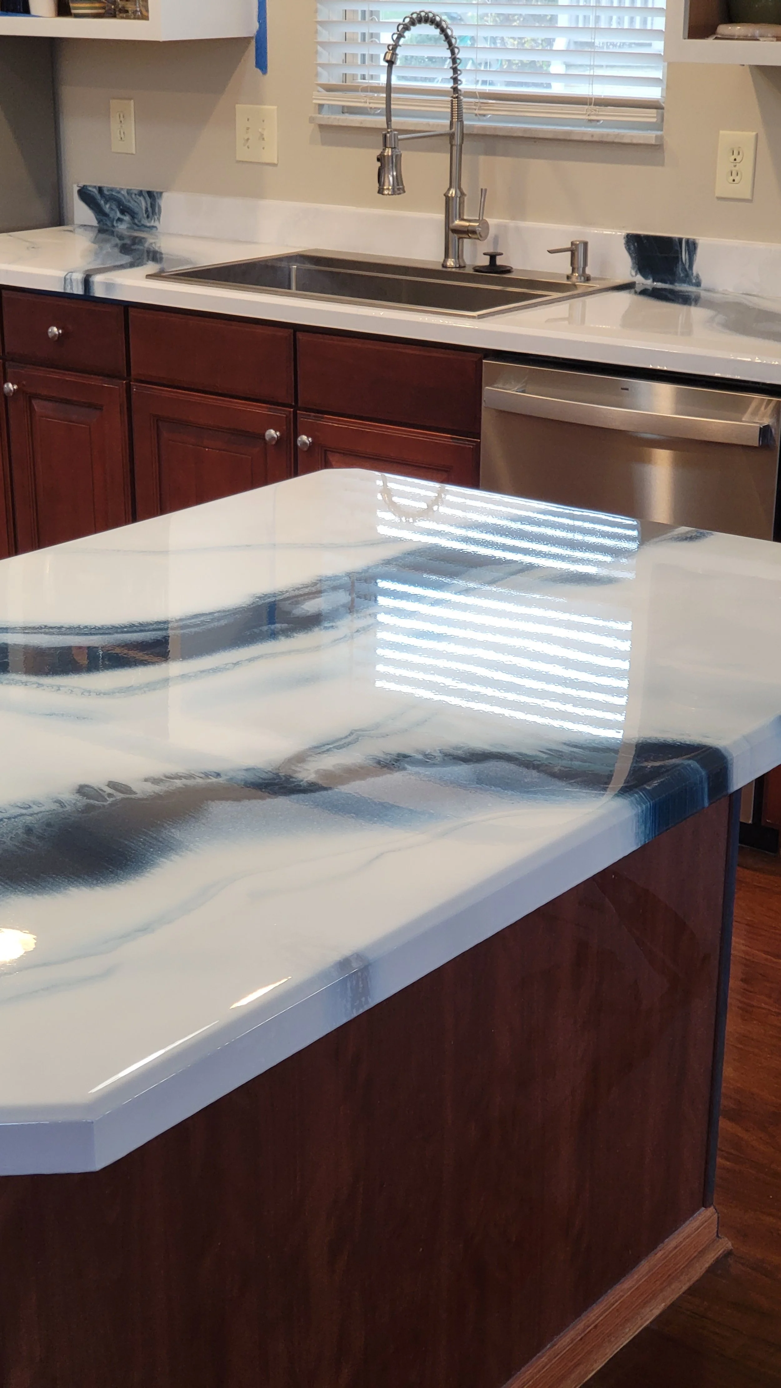 Kitchen countertop with a white and black veined marble epoxy finish, dark wooden cabinets, stainless steel sink and faucet, window with blinds, and wall outlets.