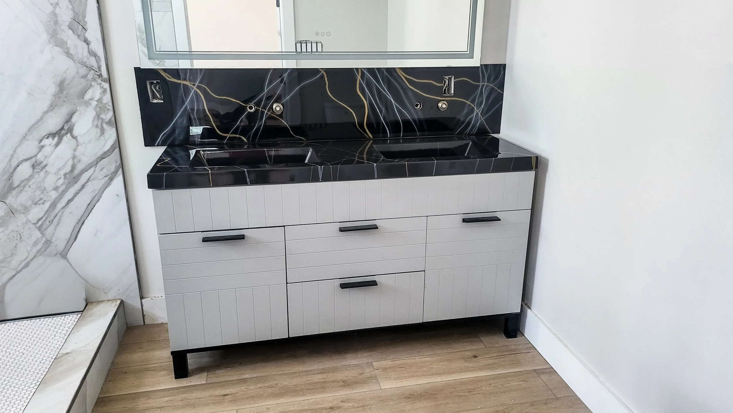 A modern bathroom vanity with two black sinks, a large mirror, black handles on drawers, and a black epoxy marble countertop with gold and white veining.