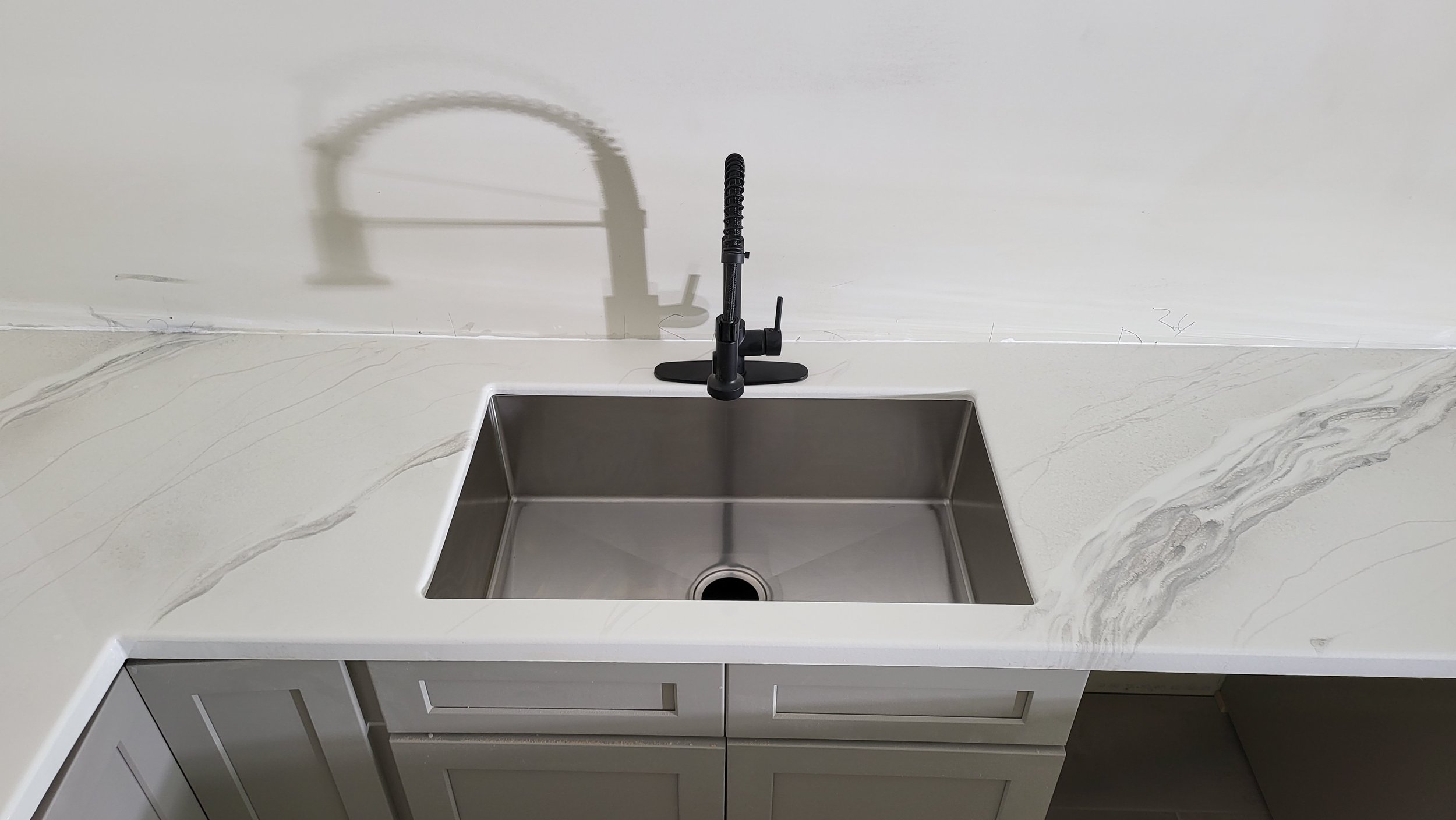 A modern kitchen sink with a black pull-down faucet mounted on a white epoxy marble countertop, undermounted stainless steel basin, with grey cabinet below.