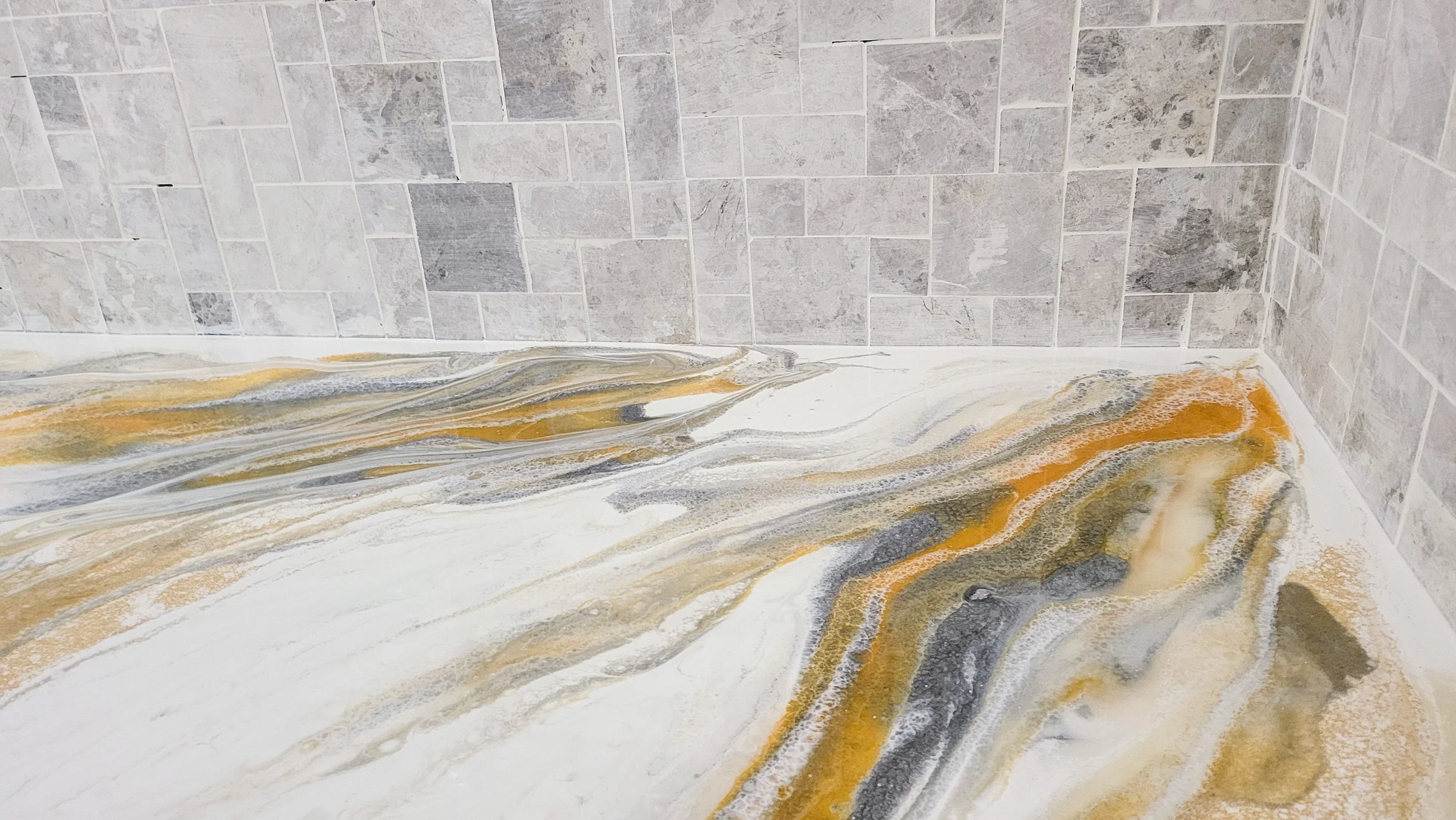 Corner of a bathroom with a tiled wall and a marble-epoxy floor in shades of white, gray, black, and gold.