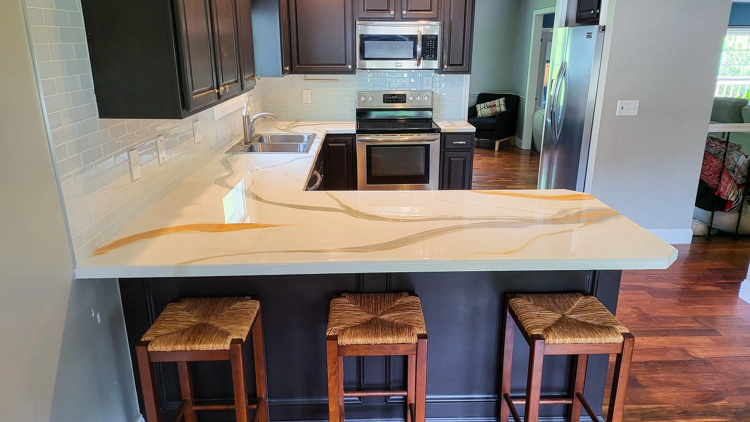 Kitchen with dark cabinets, white marble epoxy countertop with orange and brown veining, stainless steel appliances including oven, microwave, and refrigerator, and three wooden bar stools with woven seats