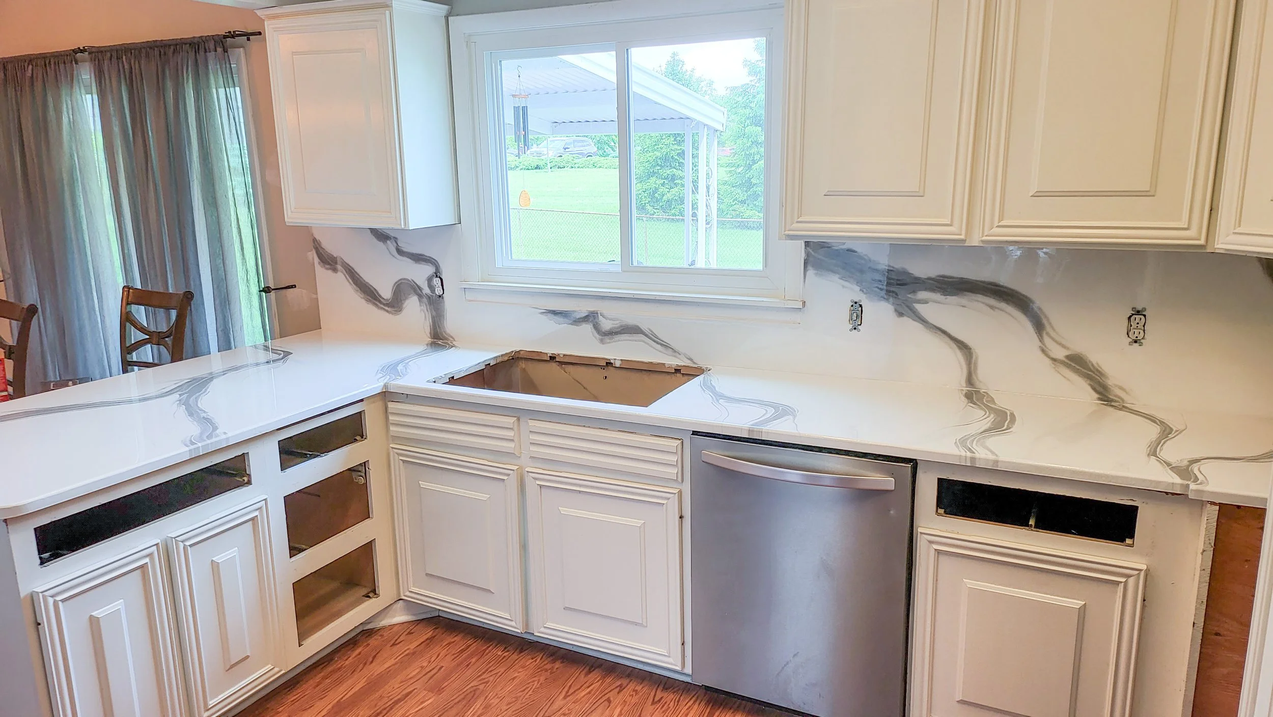 Kitchen with white cabinets and marble epoxy countertop, epoxy backsplash with black and gray veins, open space for sink, stainless steel dishwasher, window looking out to a porch and yard.