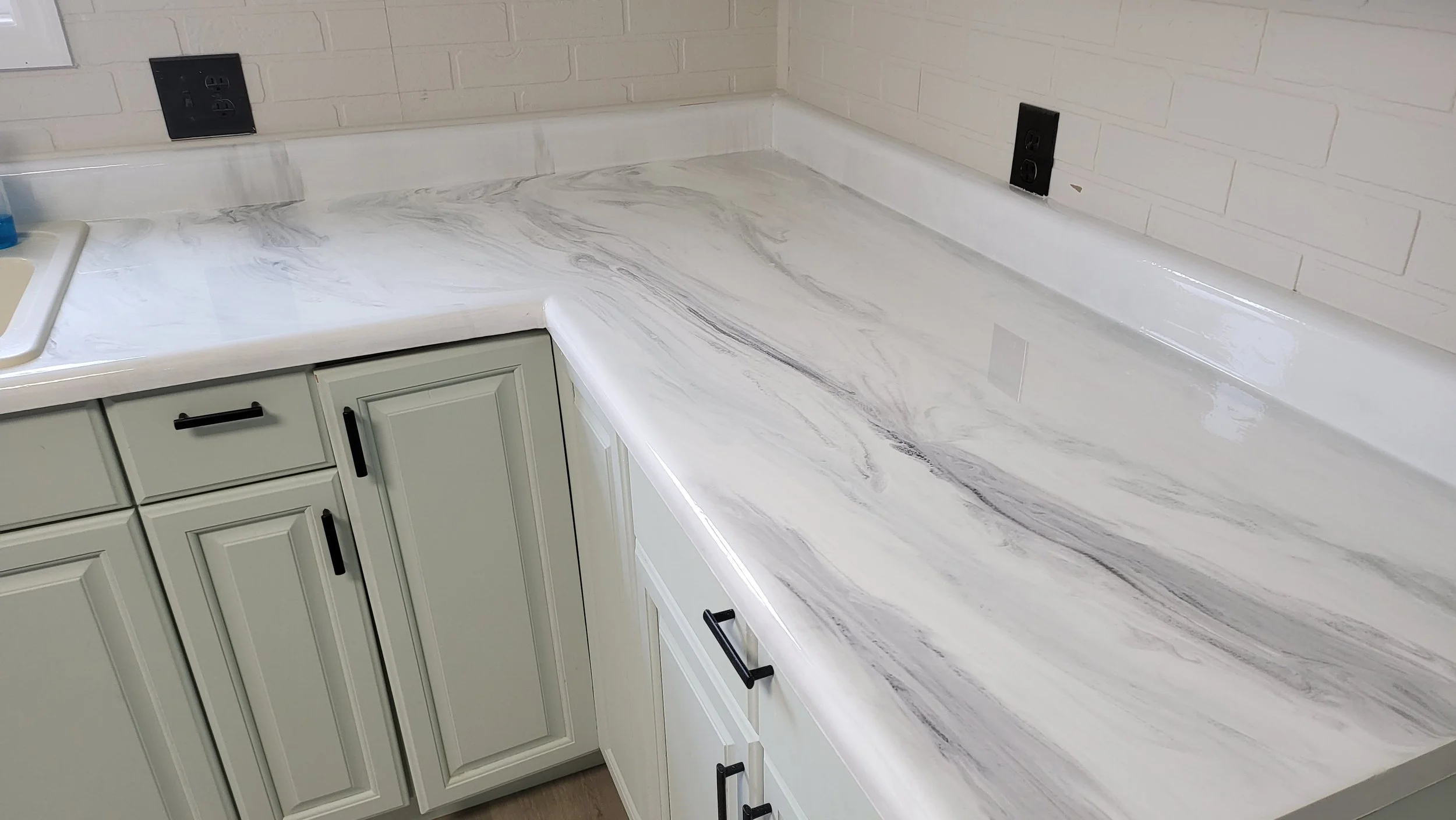 Epoxy kitchen countertop with a marble pattern, light green cabinets with black handles, white backsplash, electrical outlets, and part of a sink in the corner.
