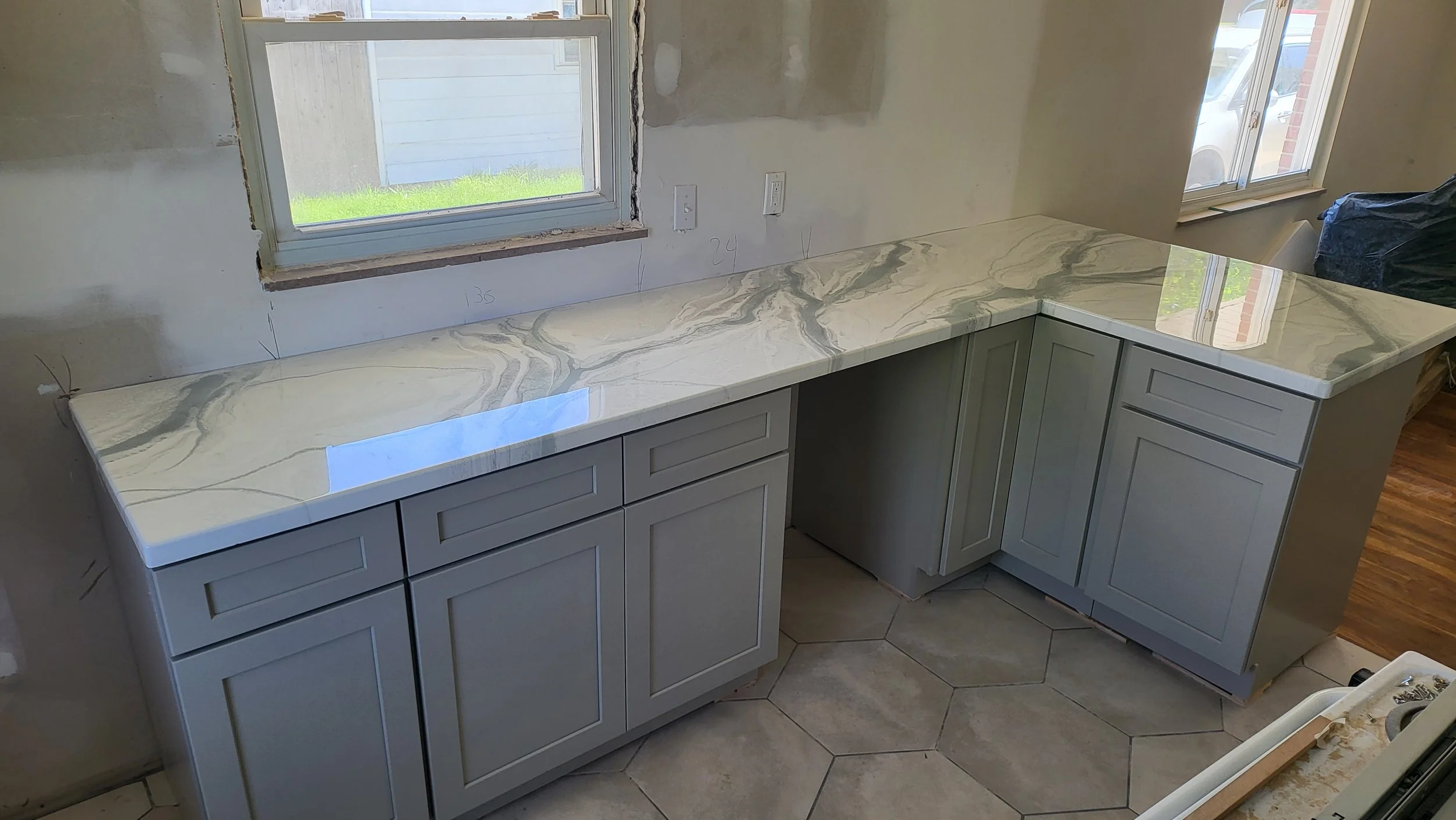 Kitchen with granite like epoxy countertop and light gray cabinets, two windows, and unfinished walls.