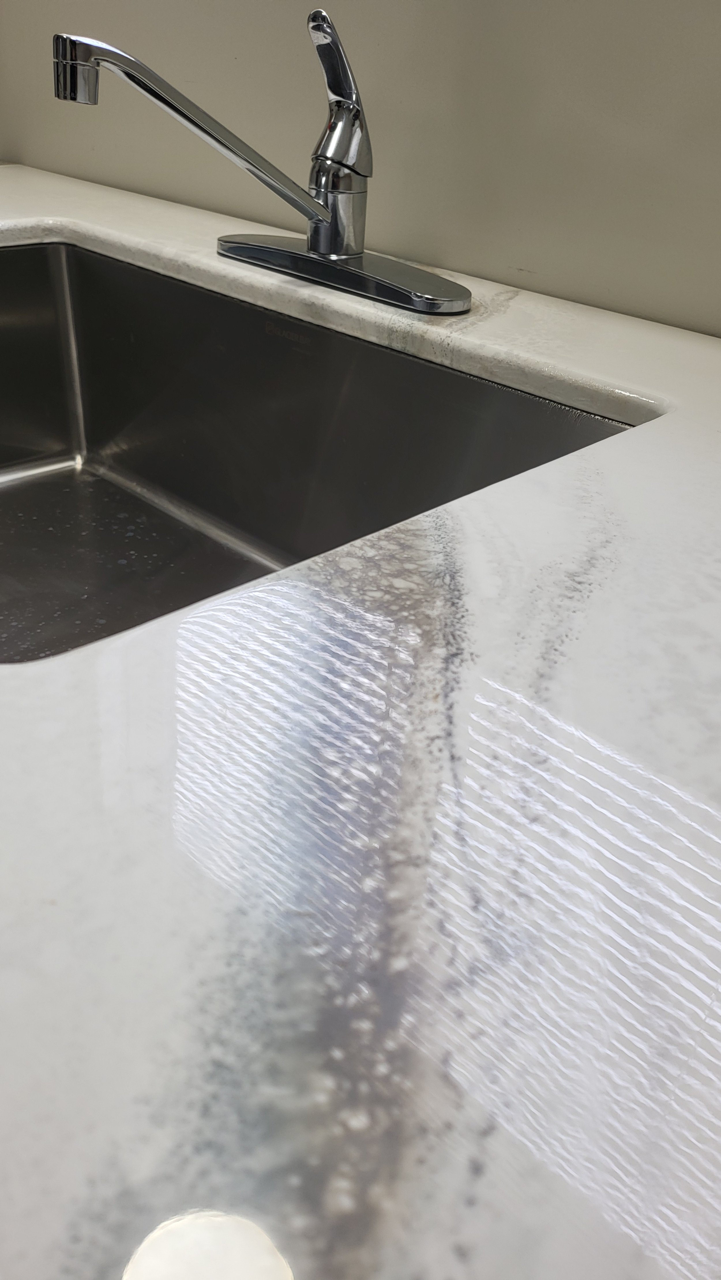A stainless steel kitchen sink with a chrome faucet on a white epoxy countertop, with water droplets inside the sink and a reflection of overhead lights on the countertop.