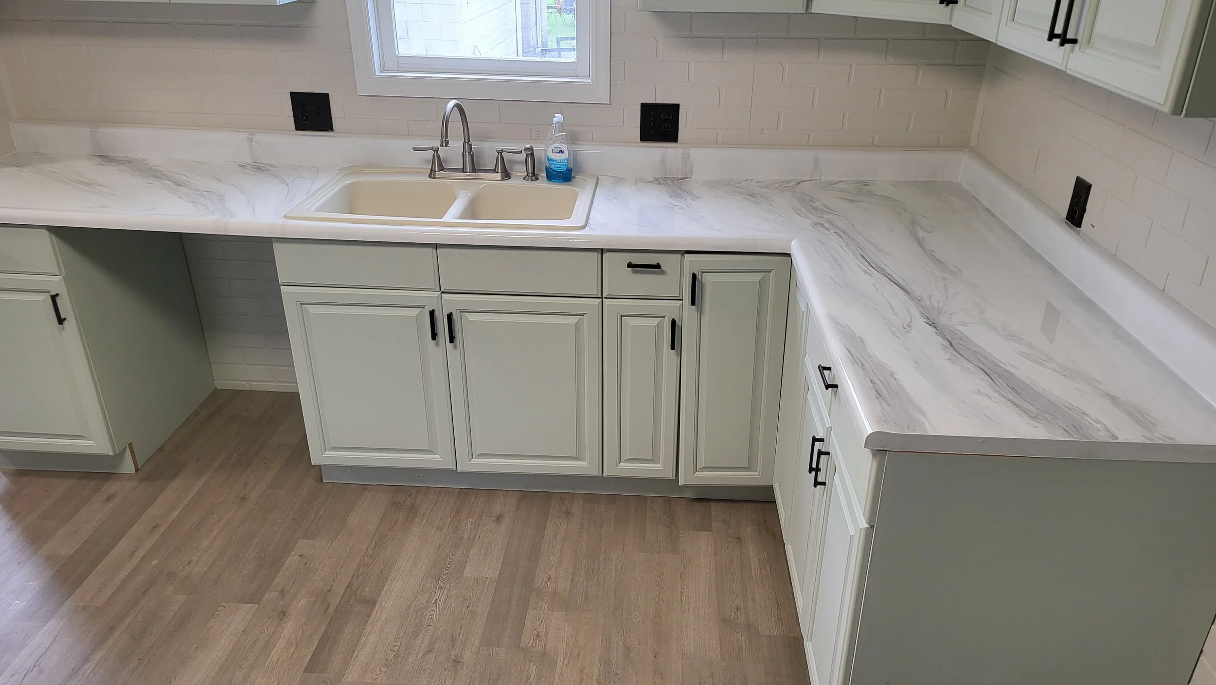 Kitchen with light green cabinetry, epoxy countertop, double sink, window above the sink, black cabinet handles, and hardwood flooring.