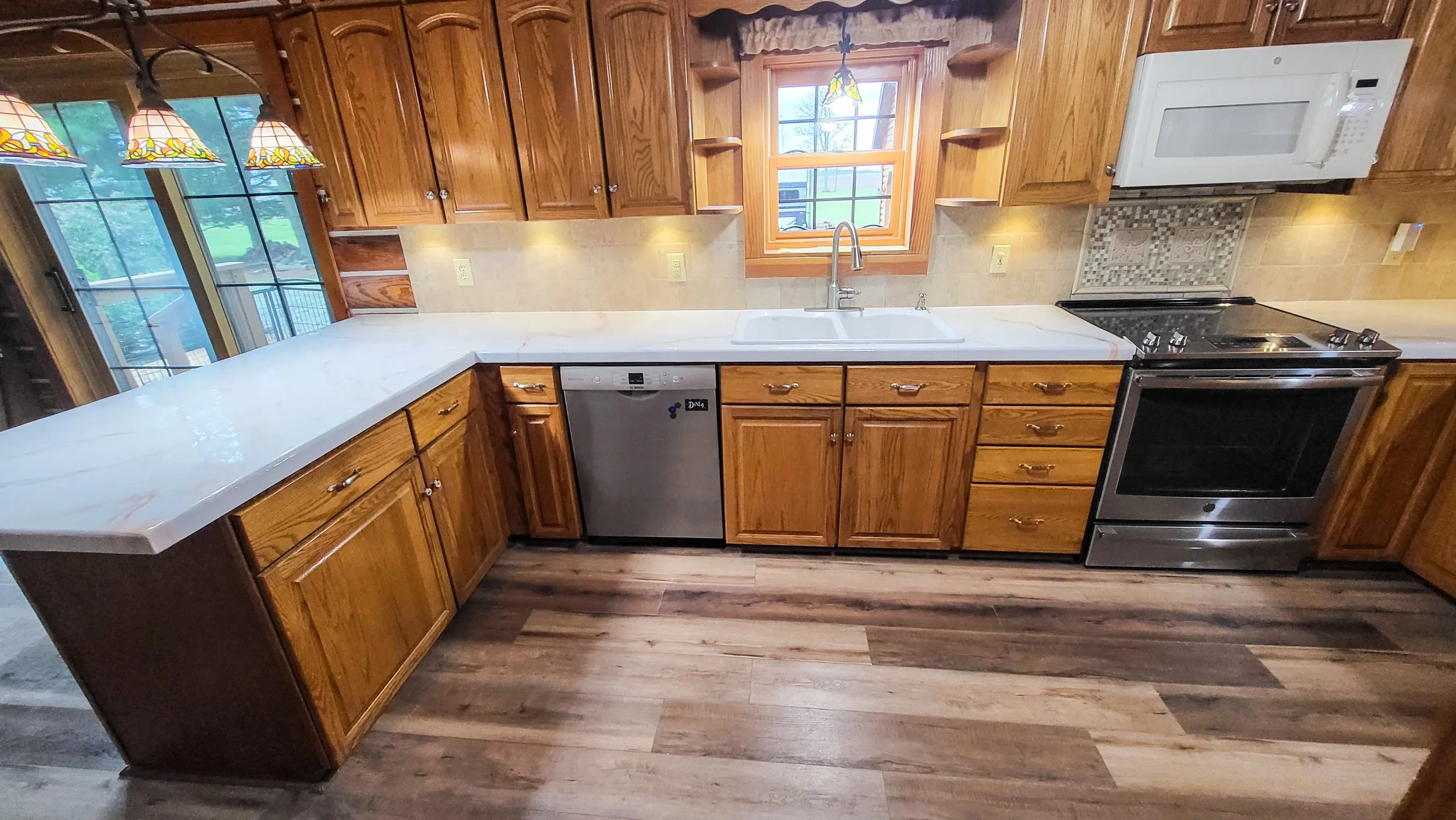 Kitchen with wooden cabinets, white epoxy countertop, stainless steel dishwasher, stove, white microwave, sink under window, tile backsplash, ceiling lights, and hardwood floor.