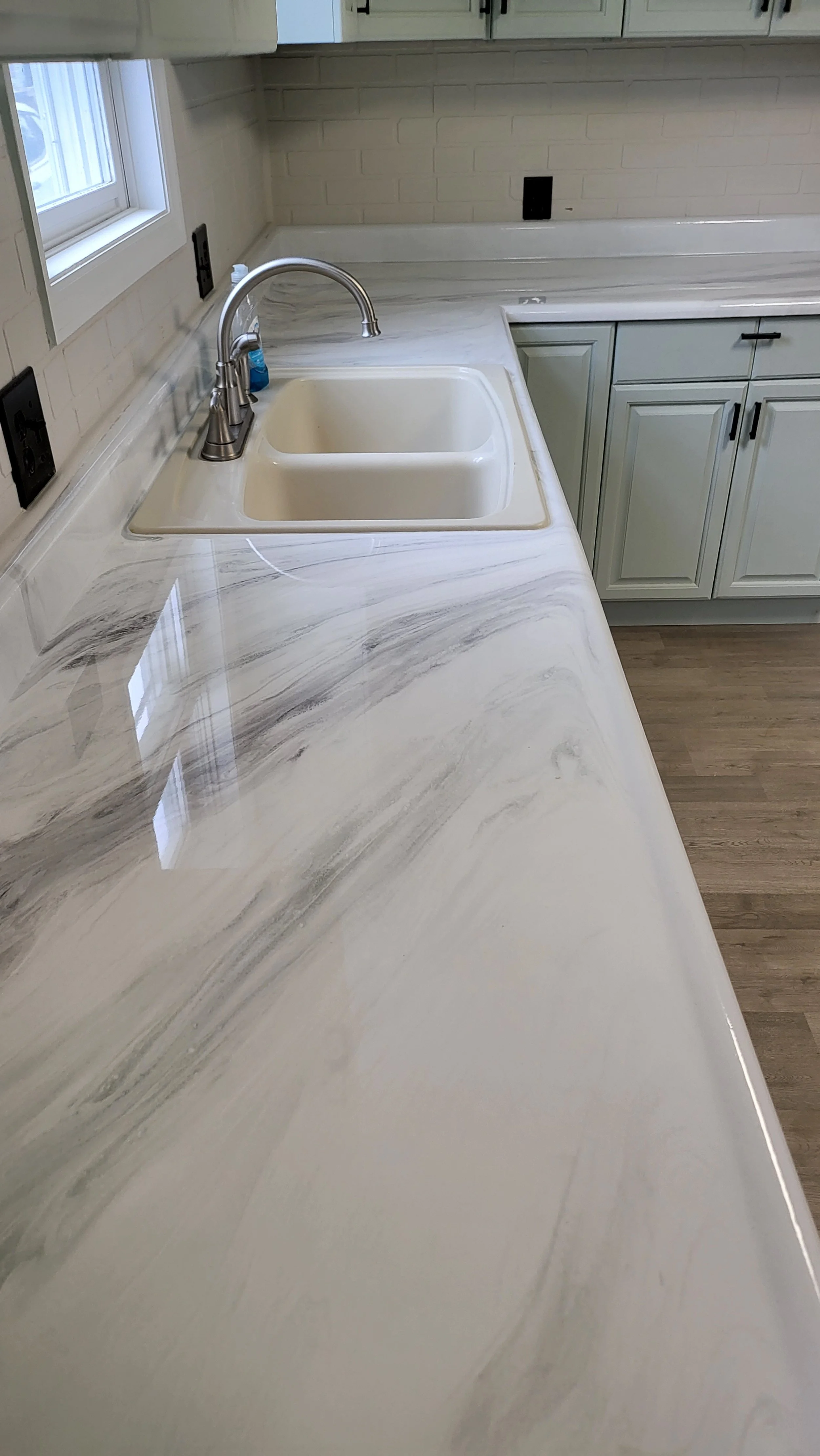 Kitchen countertop with white epoxy marble design, stainless steel sink, chrome faucet, white cabinets, and a window letting in natural light.