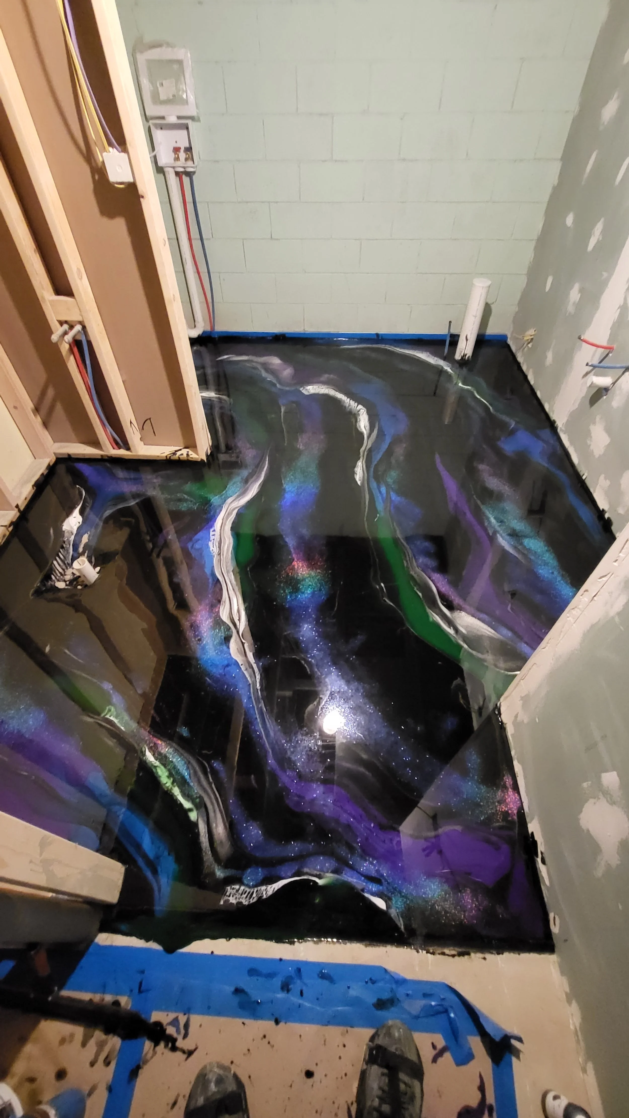 Construction site with a newly poured, glossy, black epoxy floor featuring colorful swirling patterns, seen from above with construction tools and parts of the unfinished room visible.