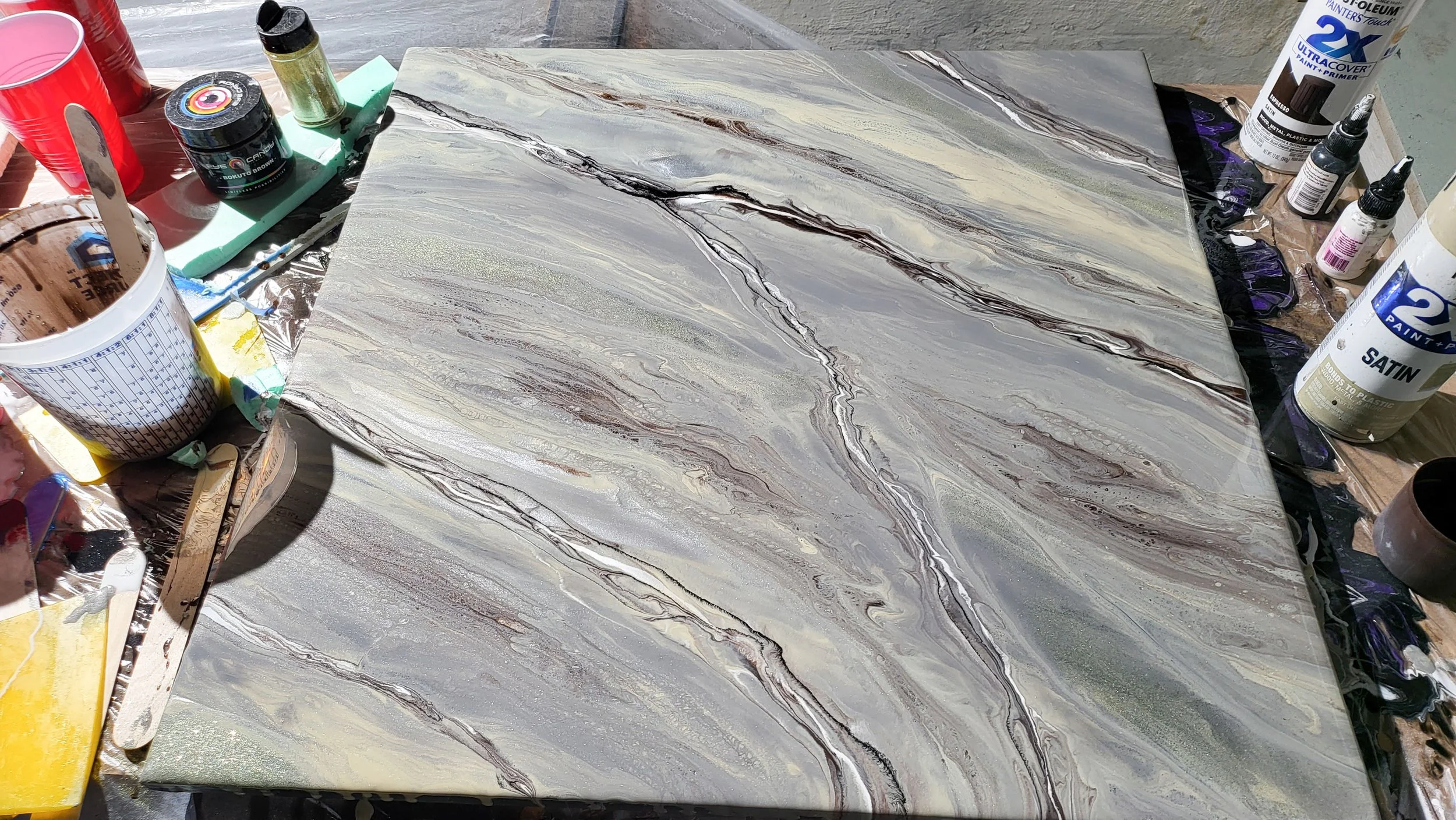 A marble-like epoxy sample board in shades of gray, white, gold, and black on a flat surface, surrounded by various painting supplies such as brushes, paints, and containers.