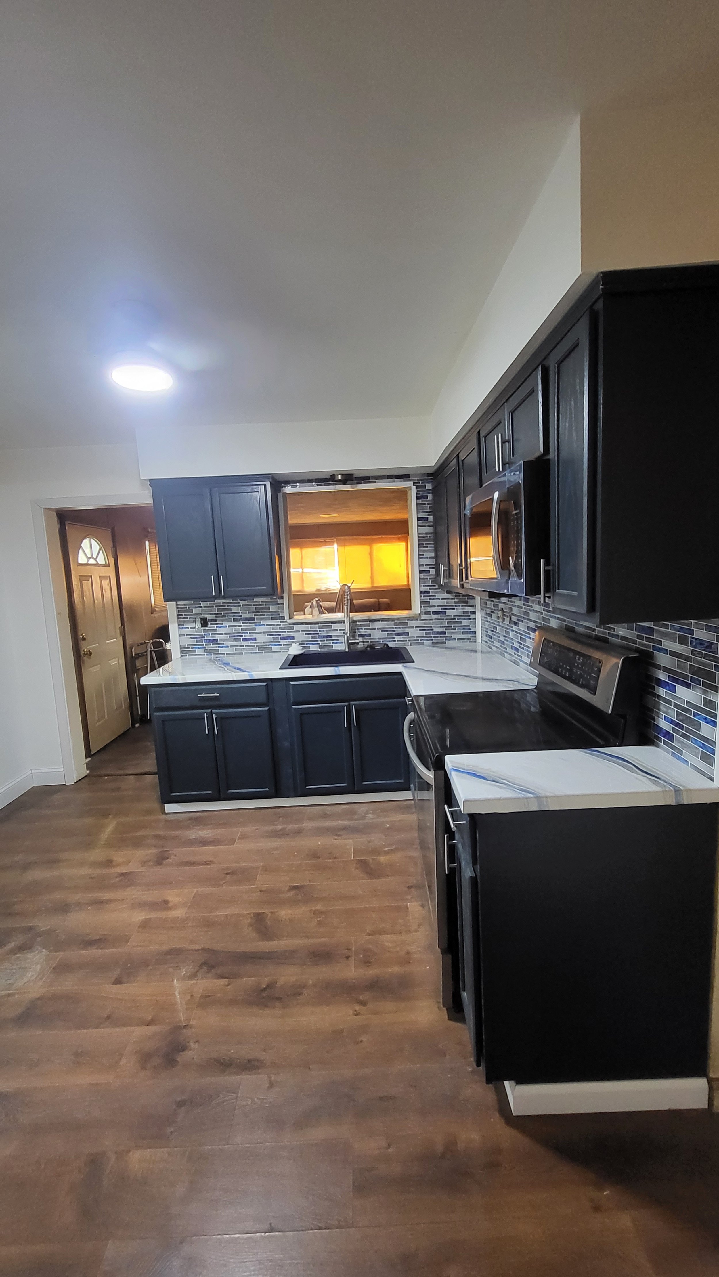 Modern kitchen with dark blue cabinets, a white epoxy countertop, a tiled backsplash, and stainless steel appliances. There is a window above the sink and a door leading outside.