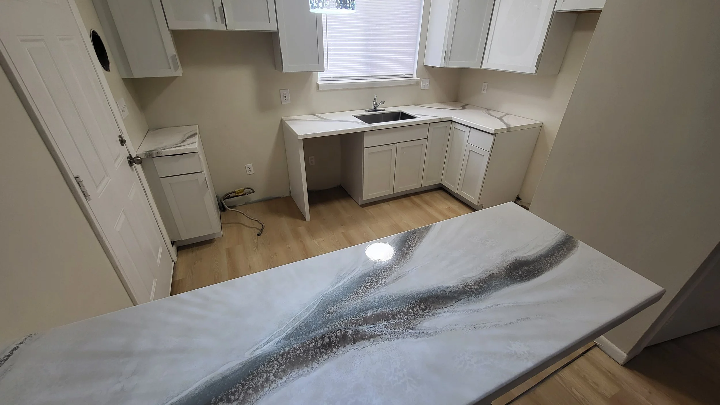 Kitchen with white cabinets and a beige epoxy countertop with gray and white swirl pattern, wood flooring, and a window with blinds.