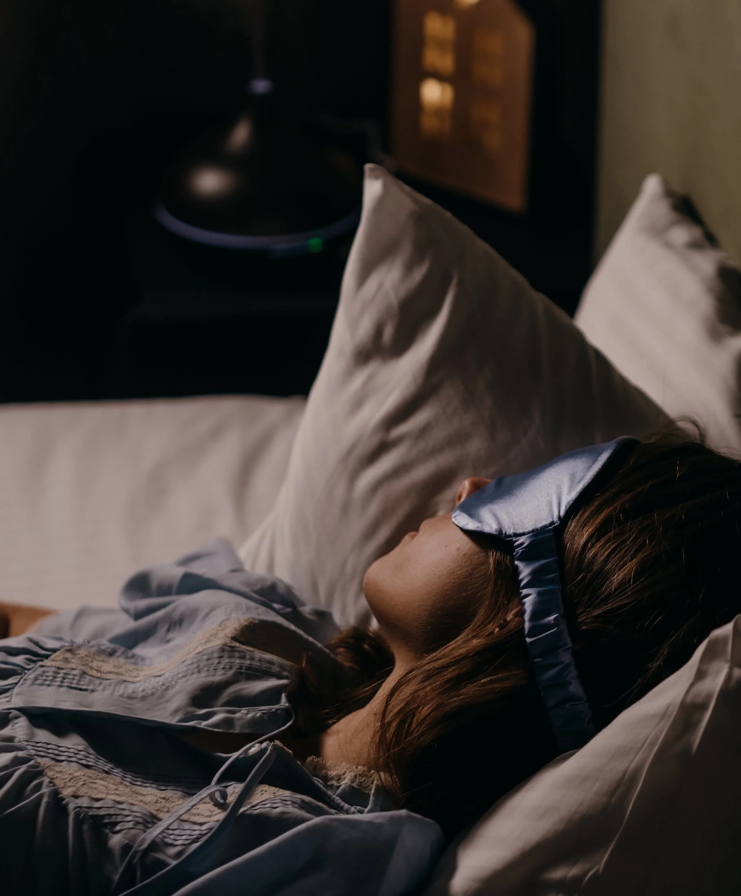 A person with long hair lying on a bed, appearing to be sleeping or resting, in a dimly lit room with a television on in the background during Ketamine Therapy Session.