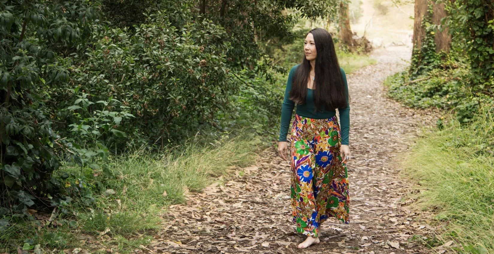 A woman walking barefoot on a leaf-covered trail in a lush, green forest, practicing mindfulness nature therapy.