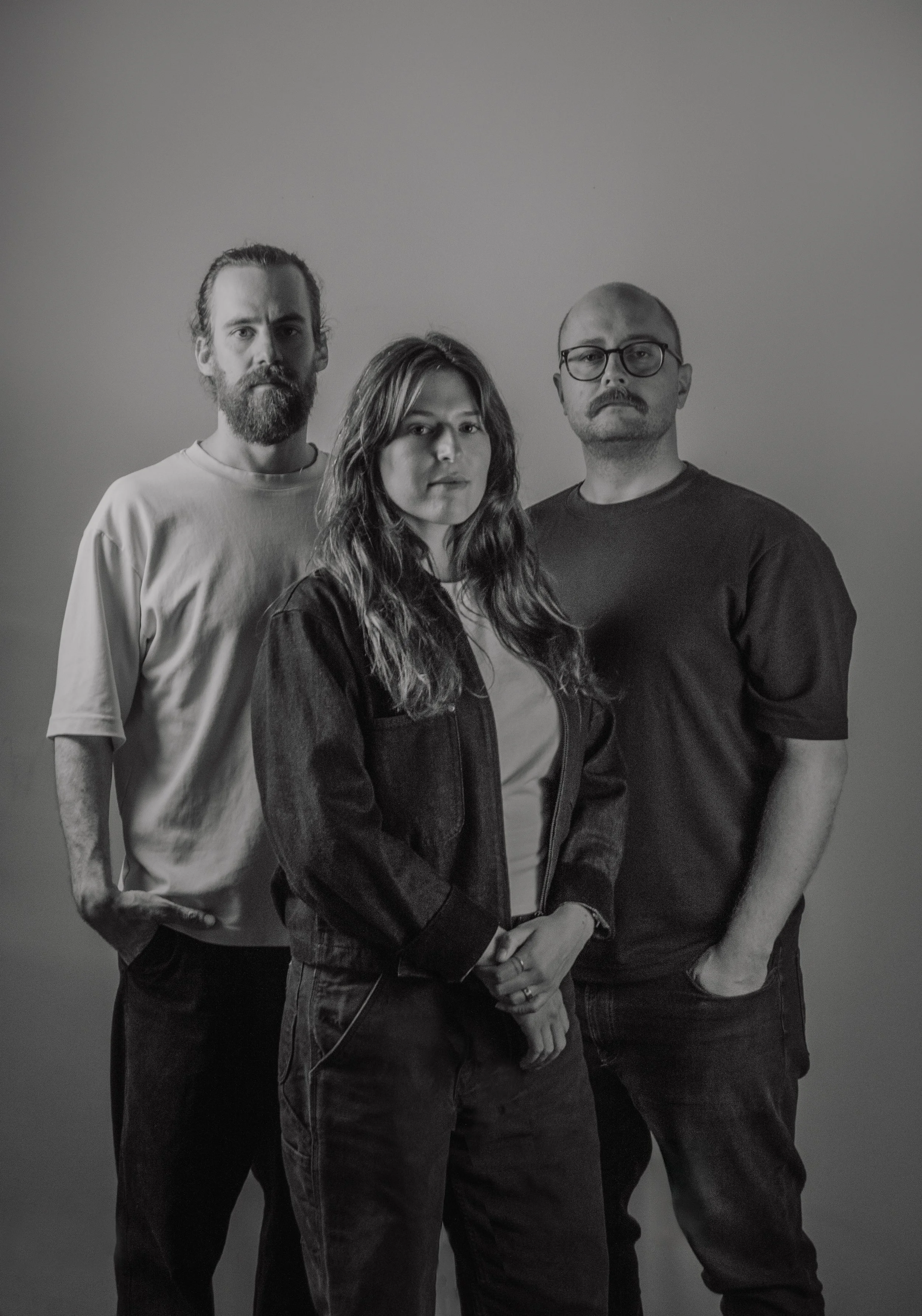Un groupe de trois personnes posant contre un fond neutre, en noir et blanc. Un homme avec des cheveux longs et une barbe à gauche, une femme aux cheveux ondulés au centre, un homme avec des lunettes à droite.