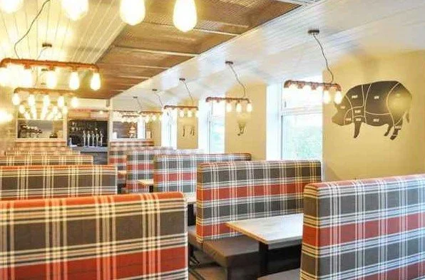 Restaurant booth seating with plaid upholstery, hanging light fixtures, and a pig silhouette mural on the wall.