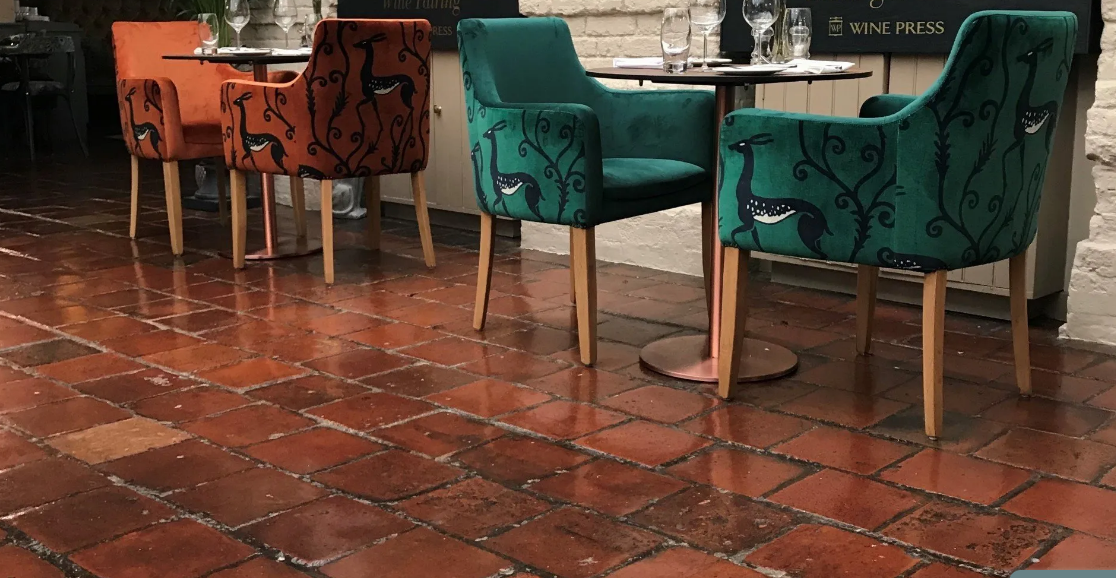 A row of four upholstered chairs with a wine-themed design, two in burnt orange and two in teal, arranged around small tables on a red brick floor in a restaurant setting.