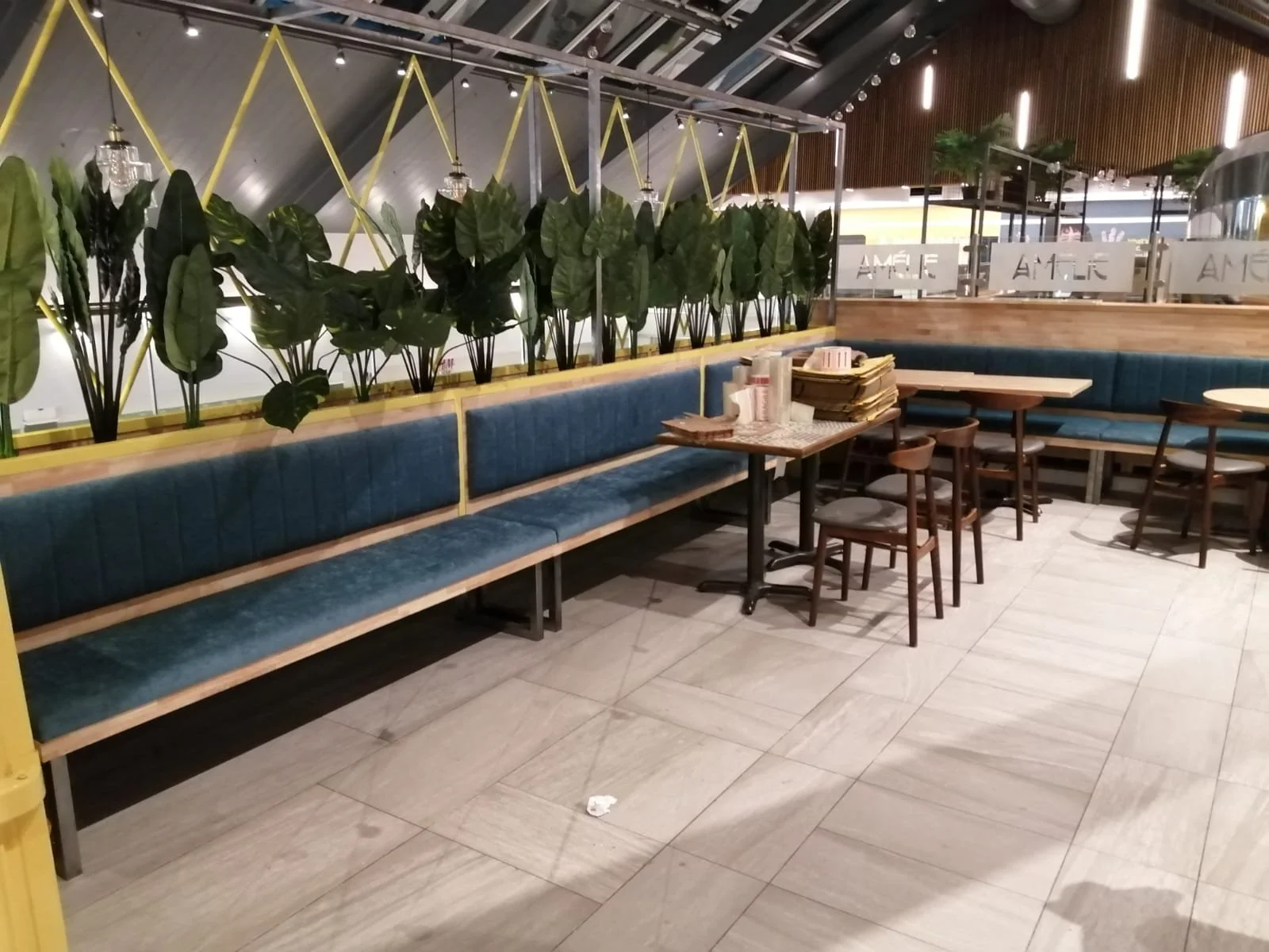 Empty restaurant seating area with blue upholstered benches, wooden tables, and chairs decorated with plants and decorative lighting