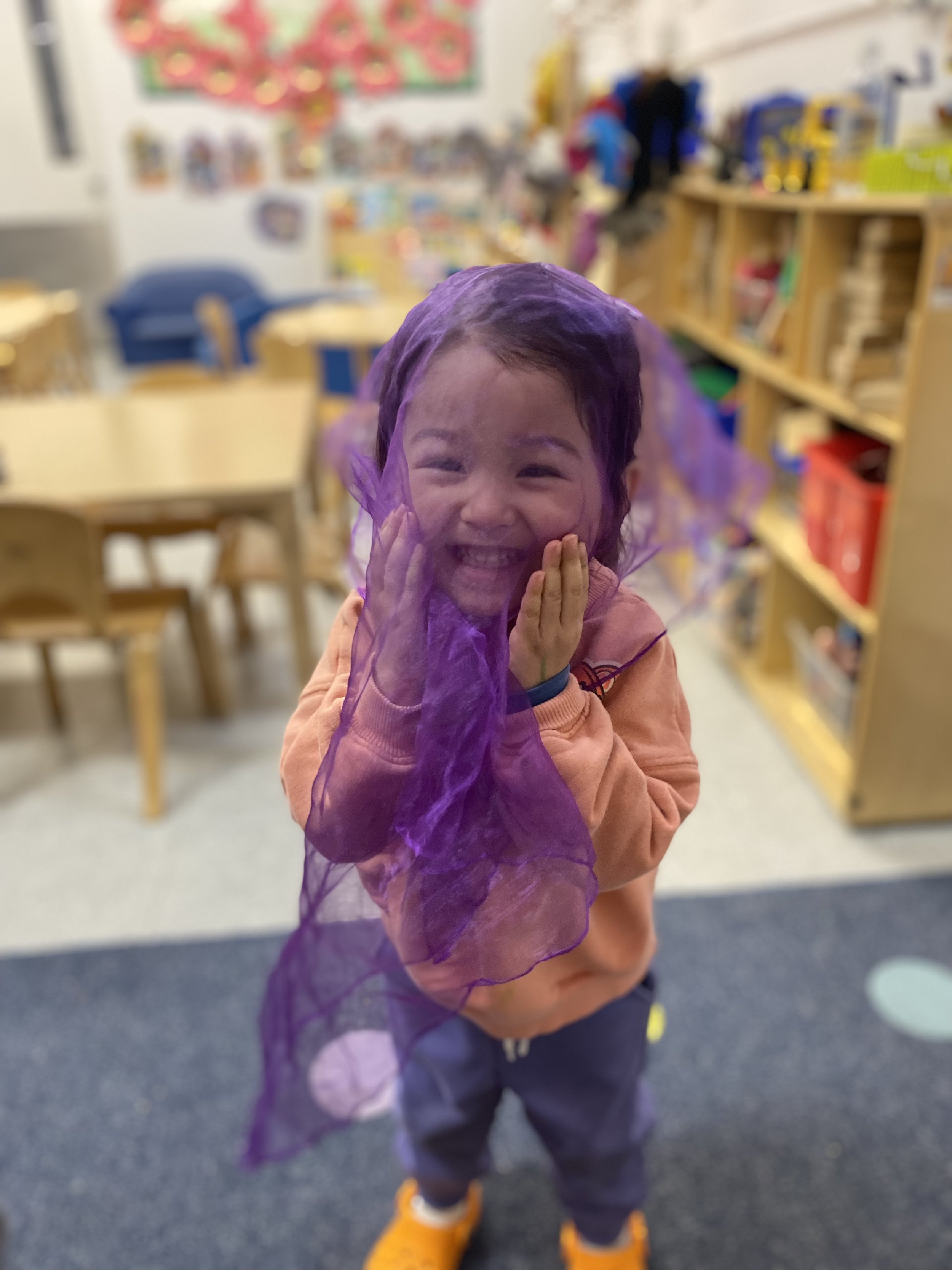Little girl with black hair and an orange shirt, plays with a purple piece of cloth in the Brownstone Brooklyn preschool with a great parent testimonial