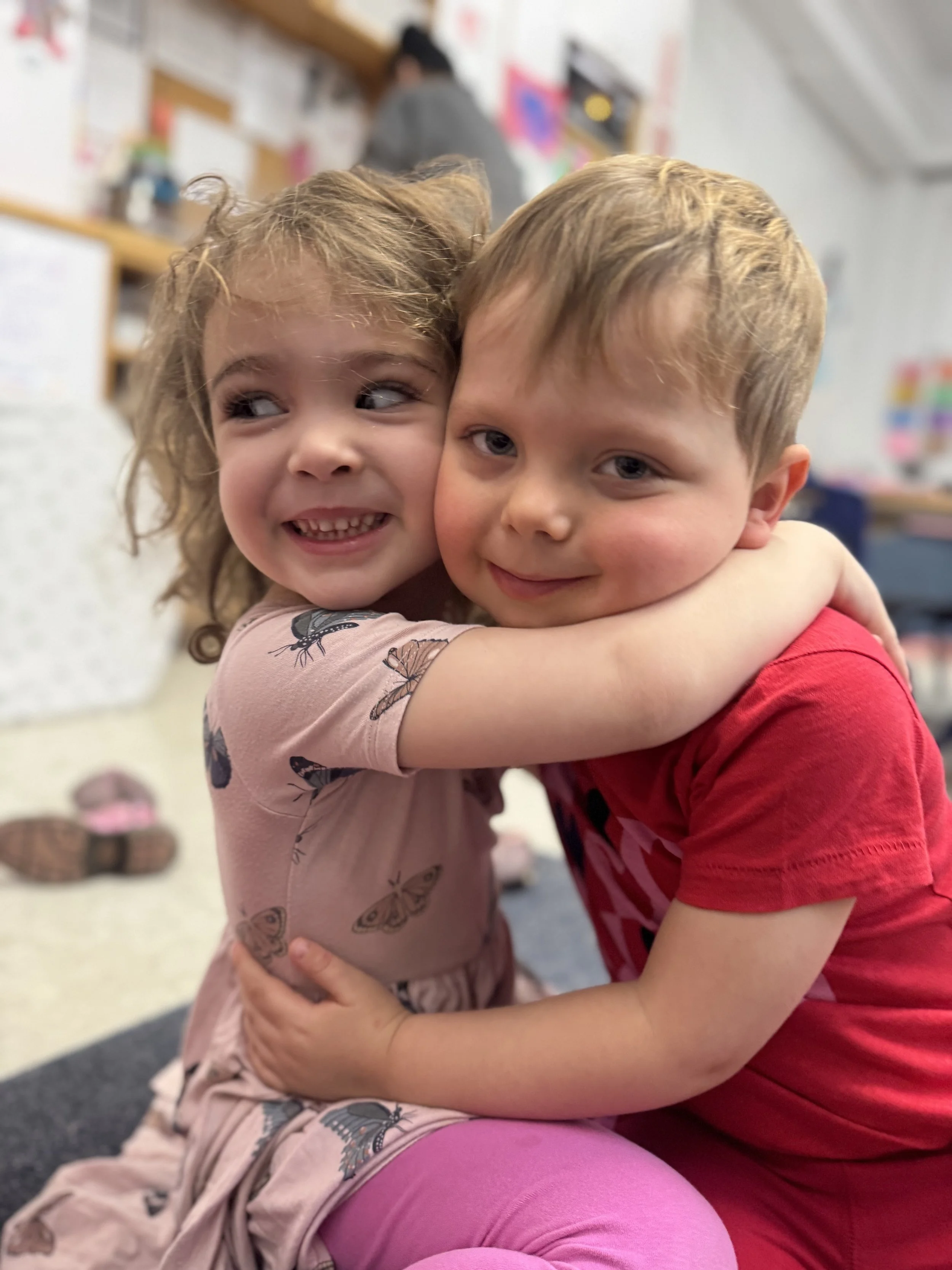 a little girl wearing pink and a little boy in red hug in a Beansprouts seeds classroom with a parent testimonial in Brooklyn preschool