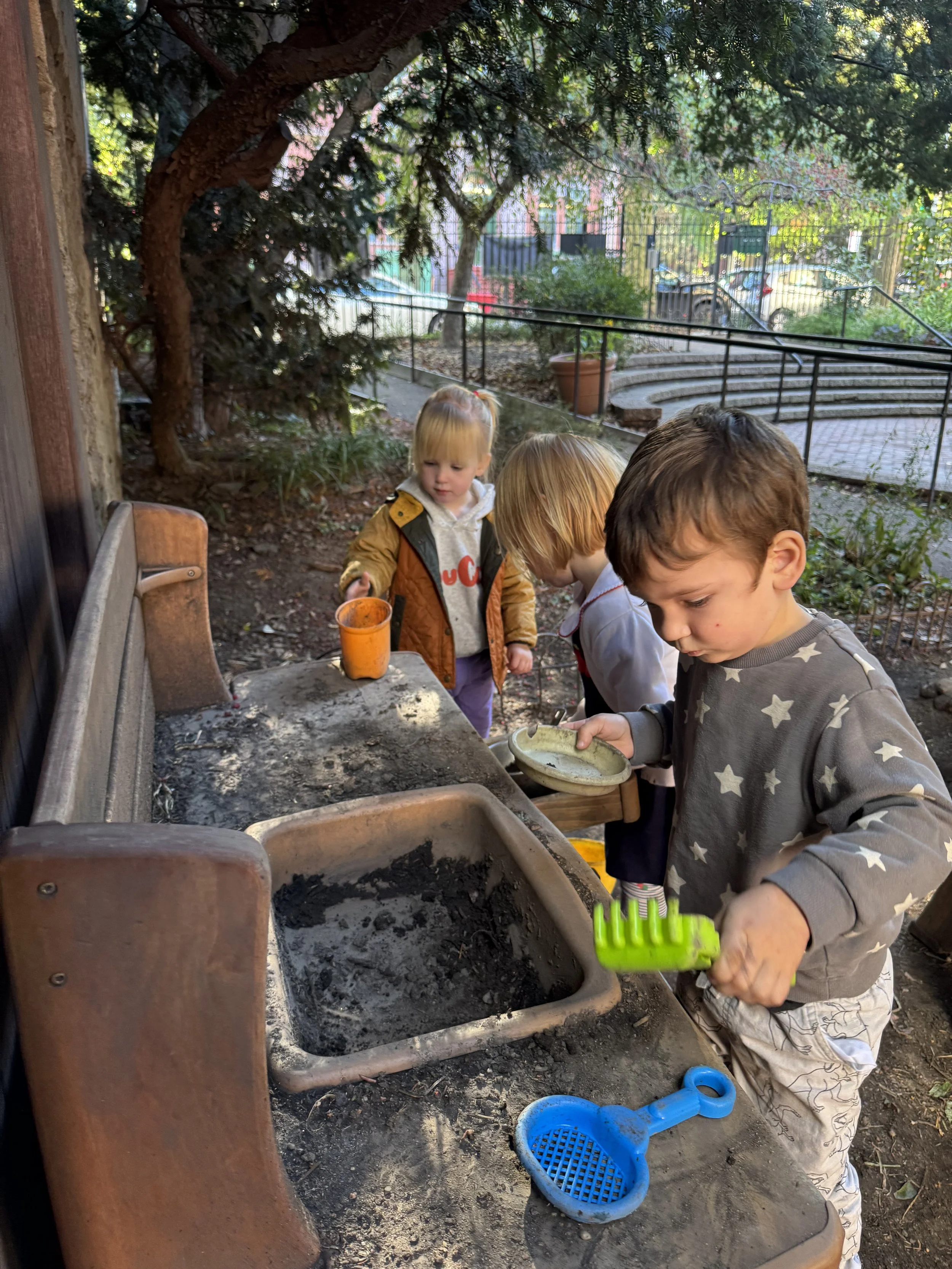 Children garden in communal outdoor area with a large sink, surrounded by trees and a park. One child is holding a green garden tool, while others are nearby in Carroll Gardens Beansprouts preschool