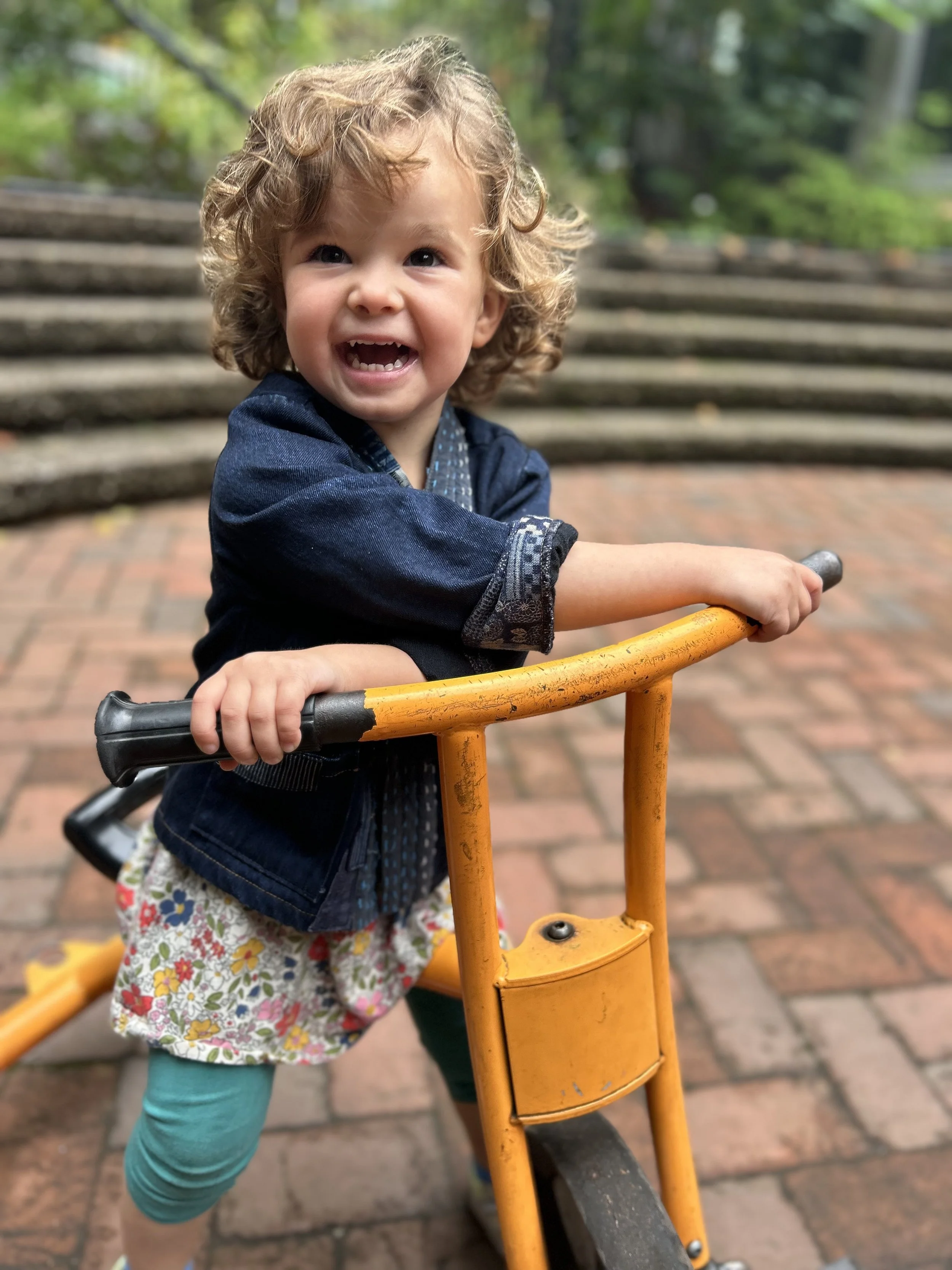 A young girl with curly hair laughing while riding a yellow tricycle on a brick pathway outdoors in Windsor Terrace Brooklyn