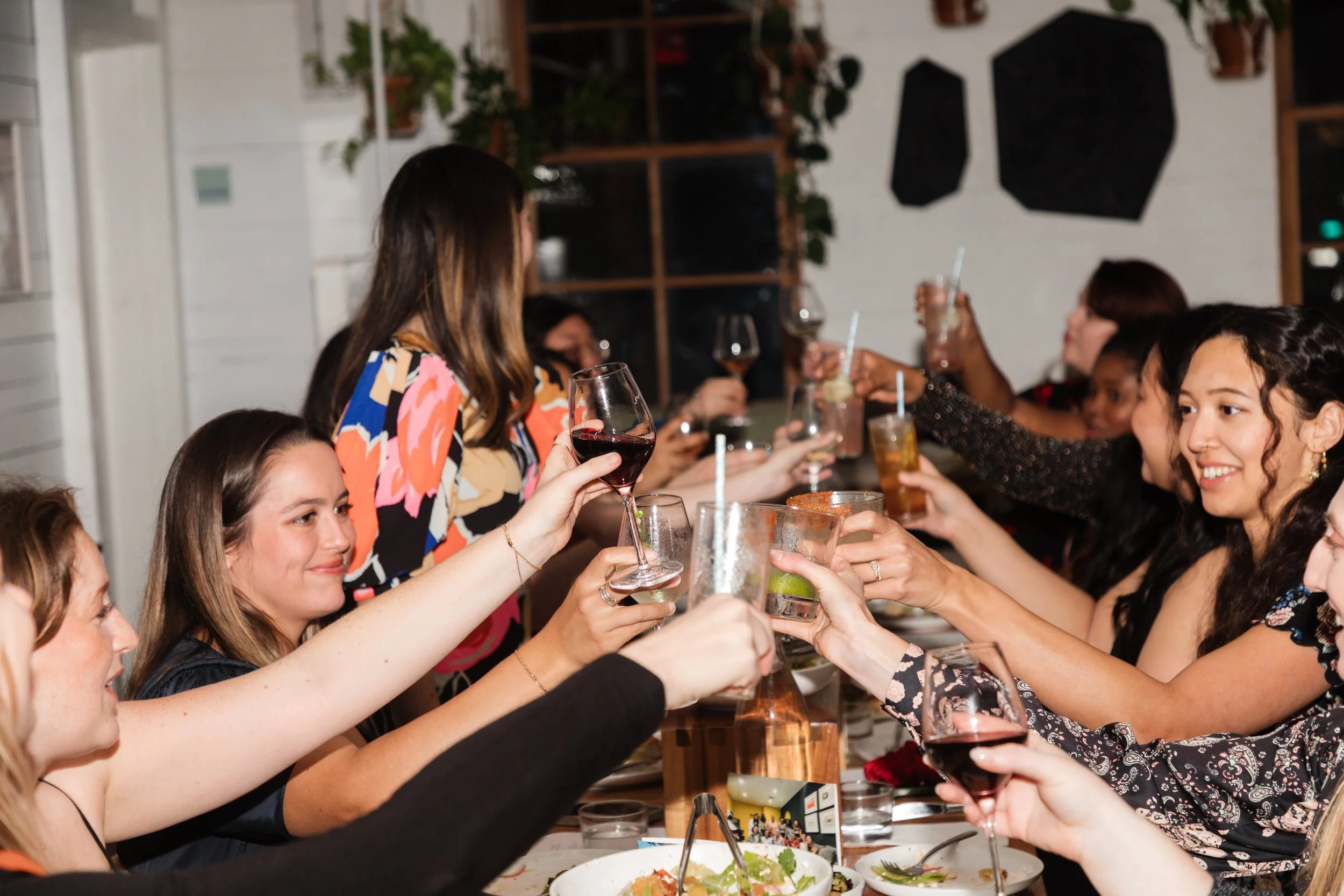 A group of women at a dinner party toasting with drinks at a long table, smiling and celebrating in a cozy, decorated indoor setting.
