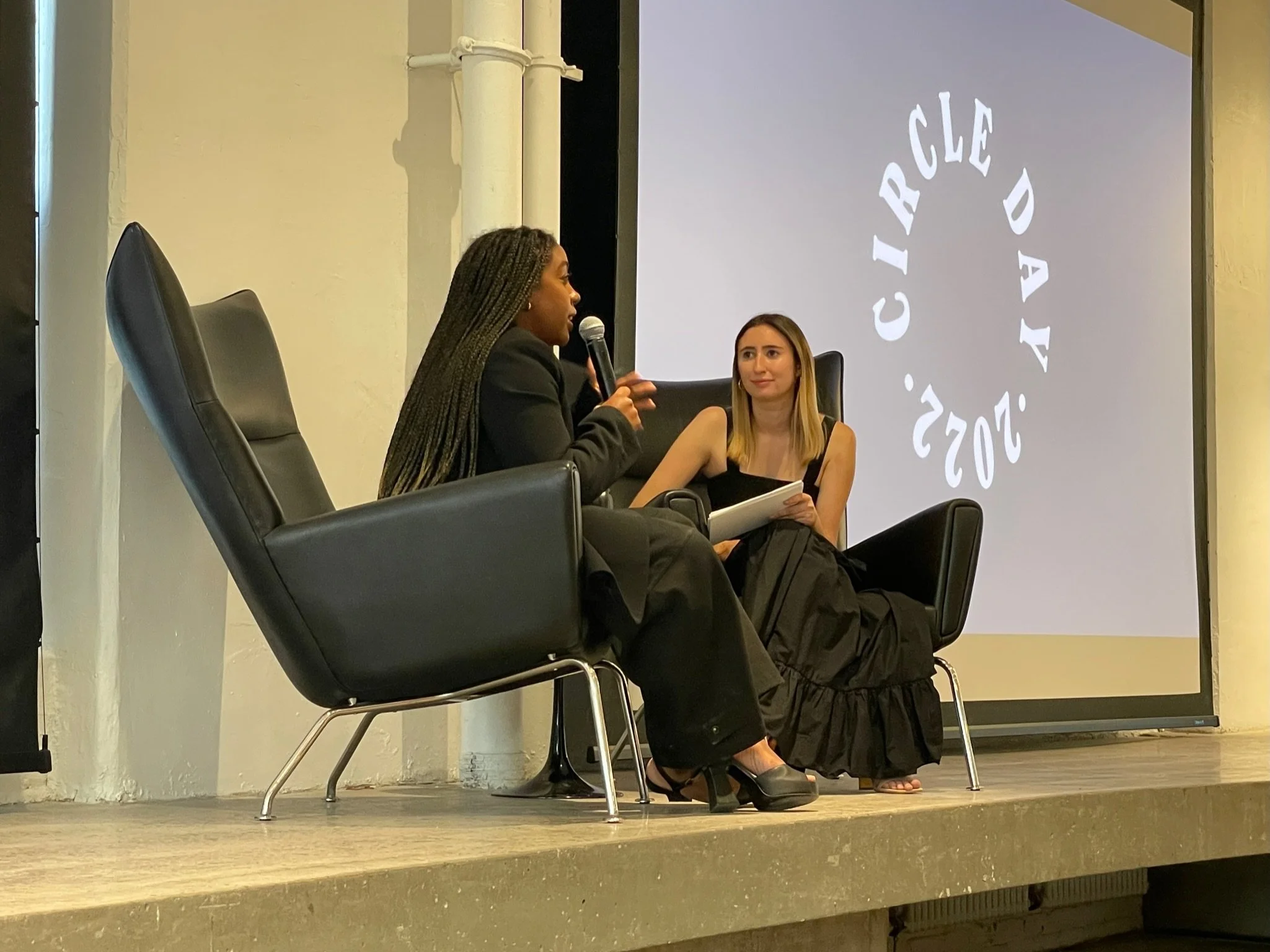 Two women sitting on a stage with black chairs, one is speaking into a microphone and the other is holding a notebook, with a large screen displaying the words 'Circle Day 2022' in the background.