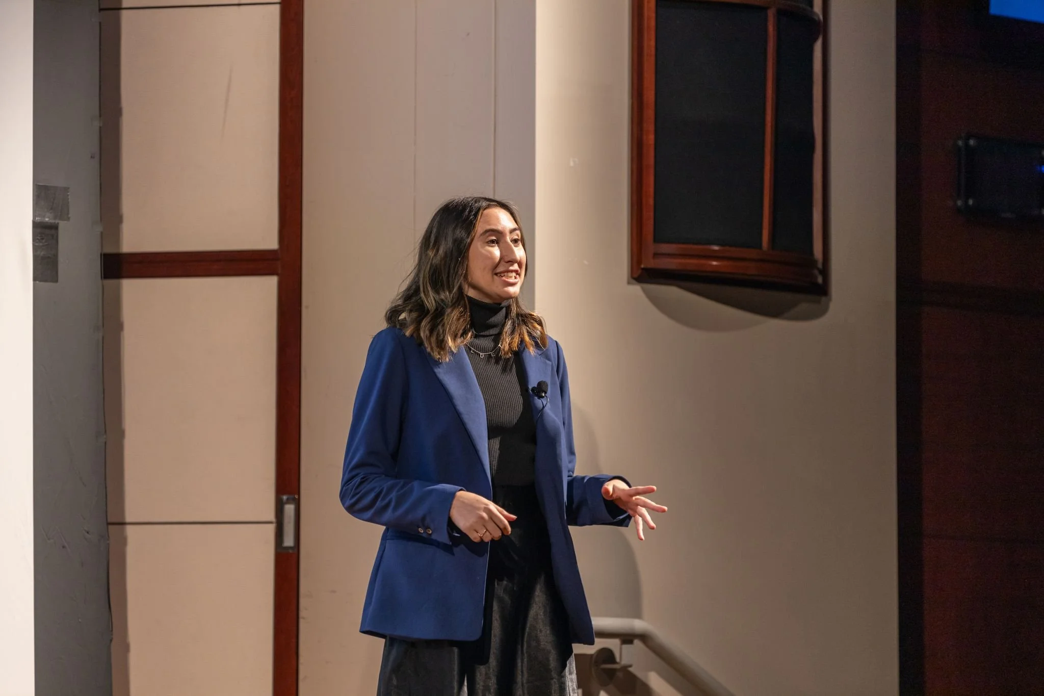 A woman with shoulder-length brown hair wearing a royal blue blazer and a black turtleneck, giving a presentation in a conference room.