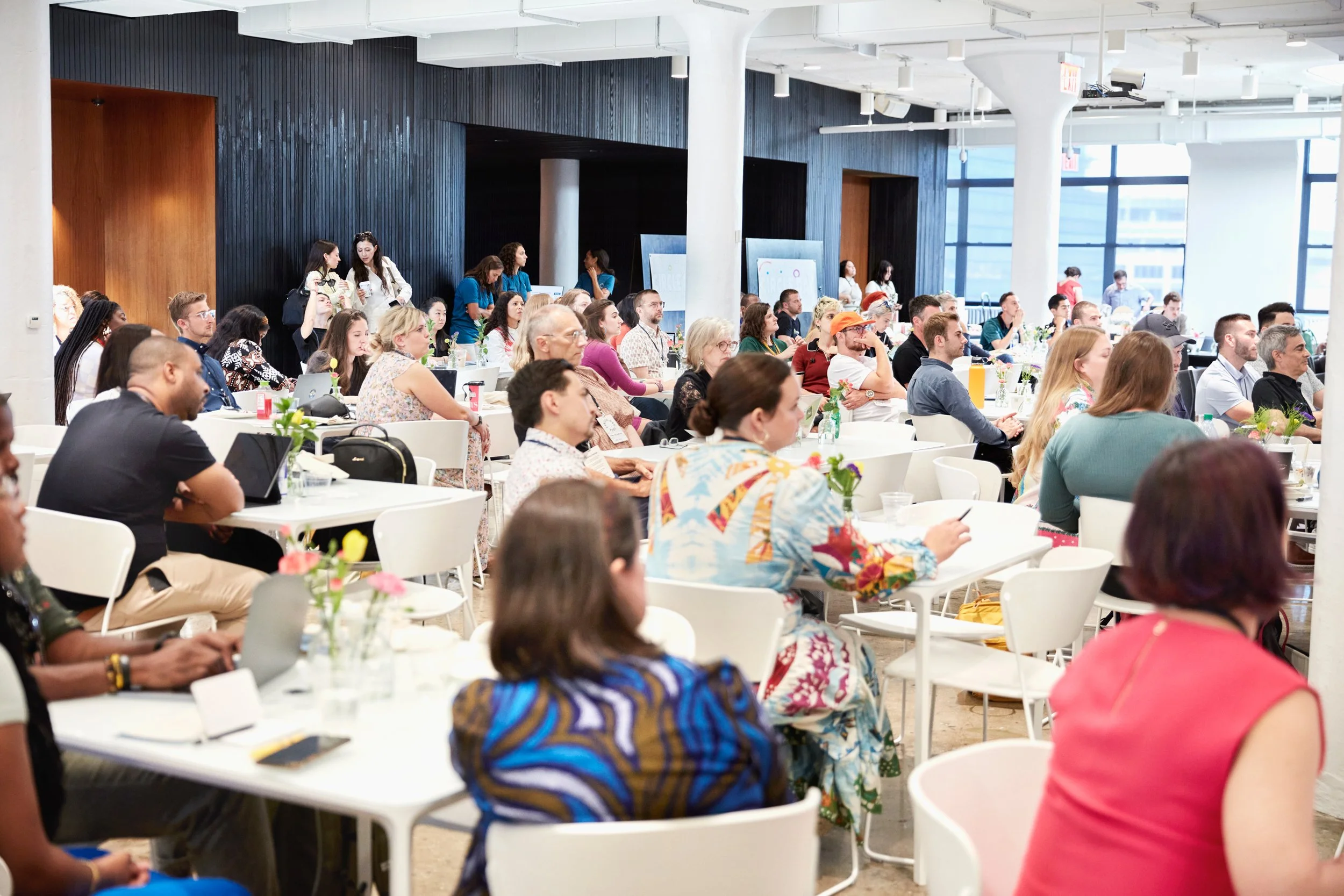 A diverse group of people sitting at white tables in a bright conference room attending a presentation or seminar.